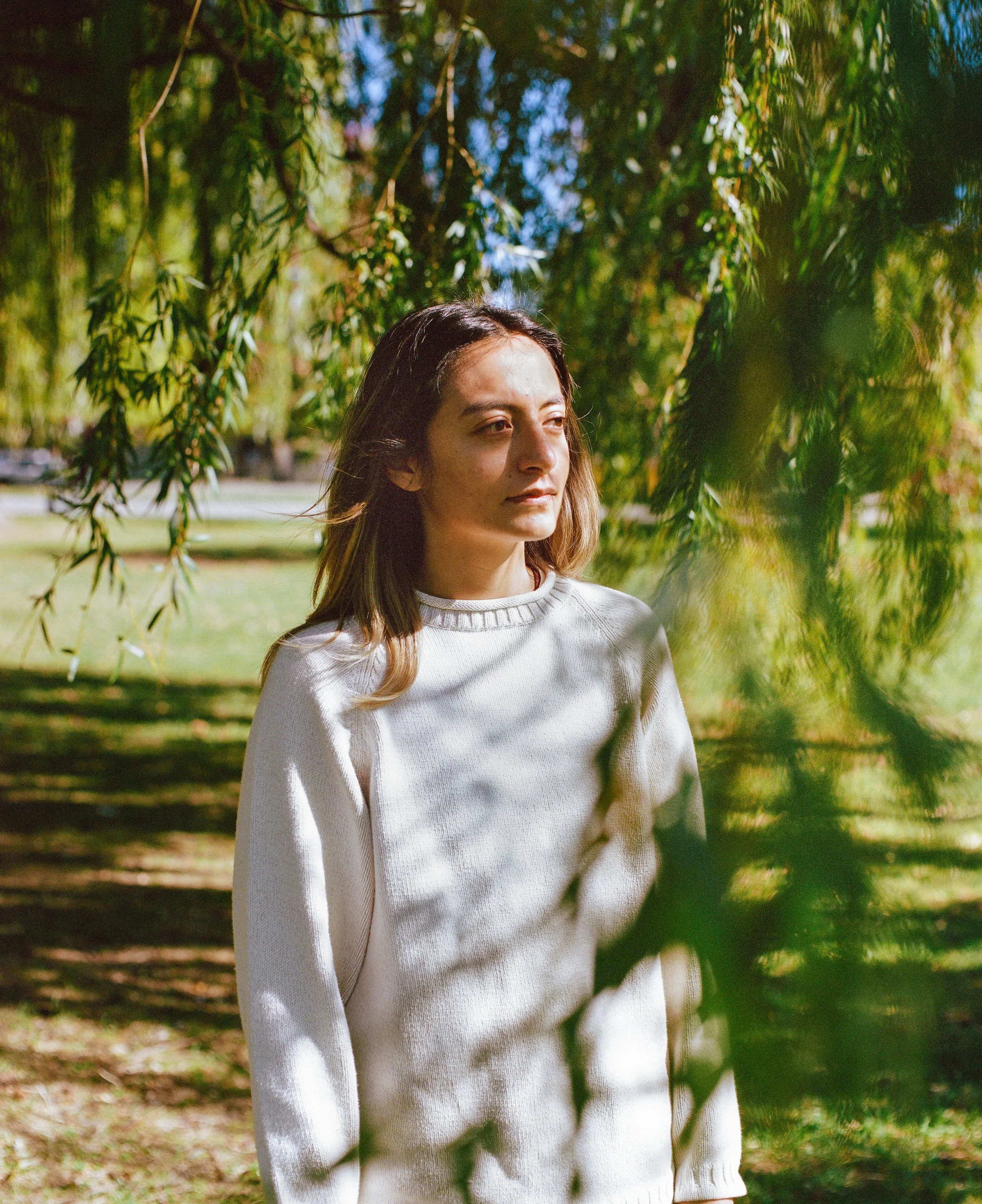 A young woman standing outdoors in a park, partially obscured by tree branches with green leaves, wearing a white sweater and looking thoughtfully into the distance.