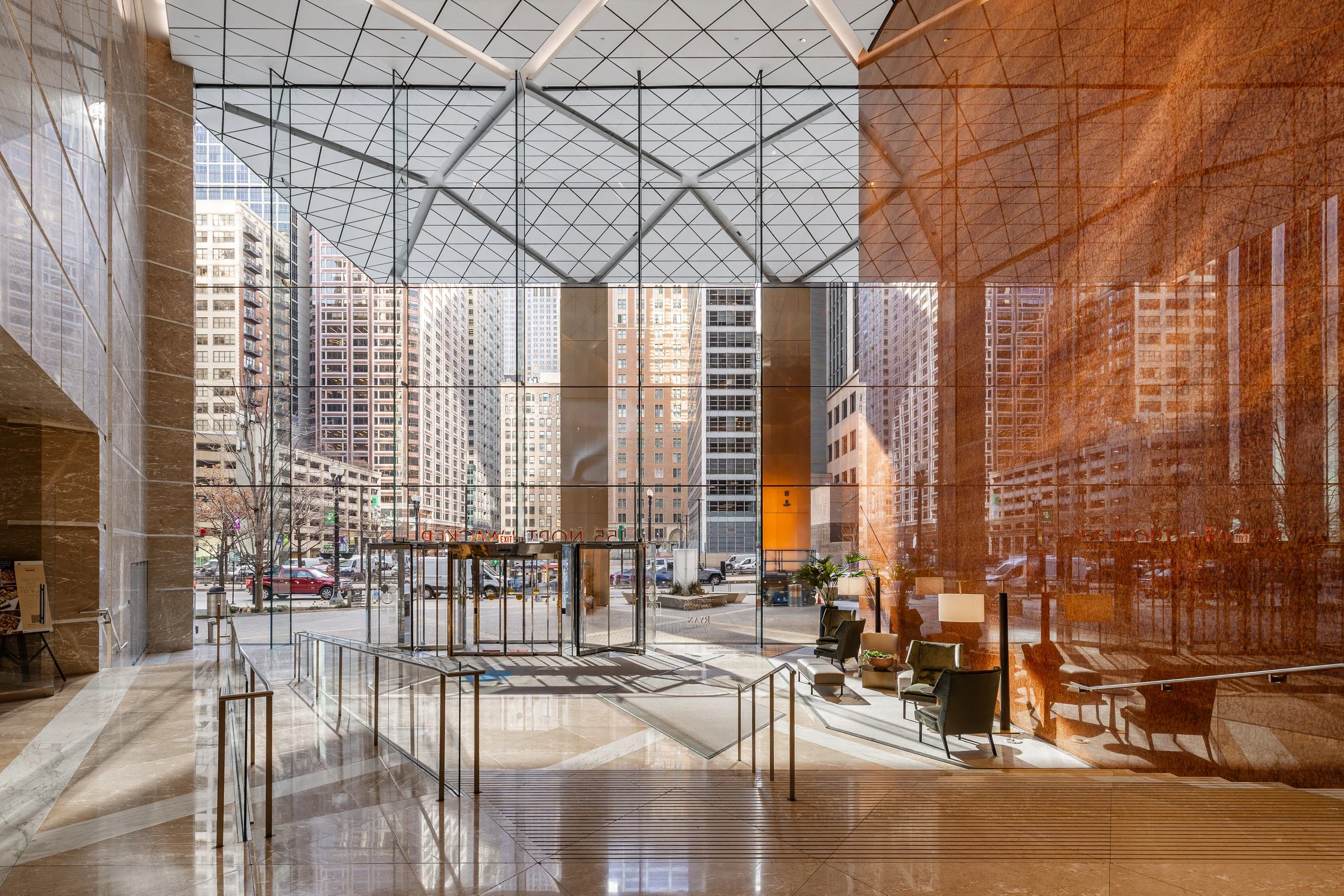 The image shows the lobby of a modern building with large glass windows. Inside, there are several chairs, a small table with plants, and a lamp. Outside, tall skyscrapers and cars are visible on the street.