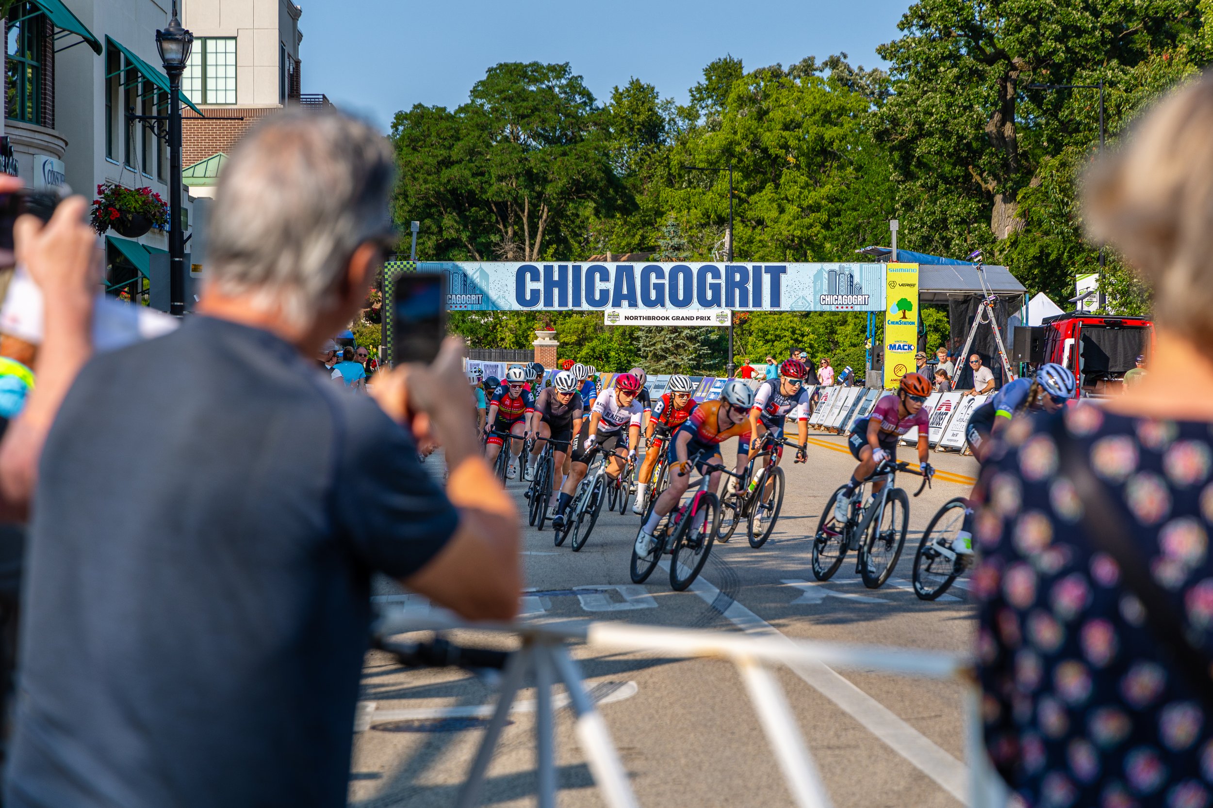 Cyclists in a race passing under a large sign that reads 'CHICAGO GRIT' during daytime with spectators taking photos and cheering.
