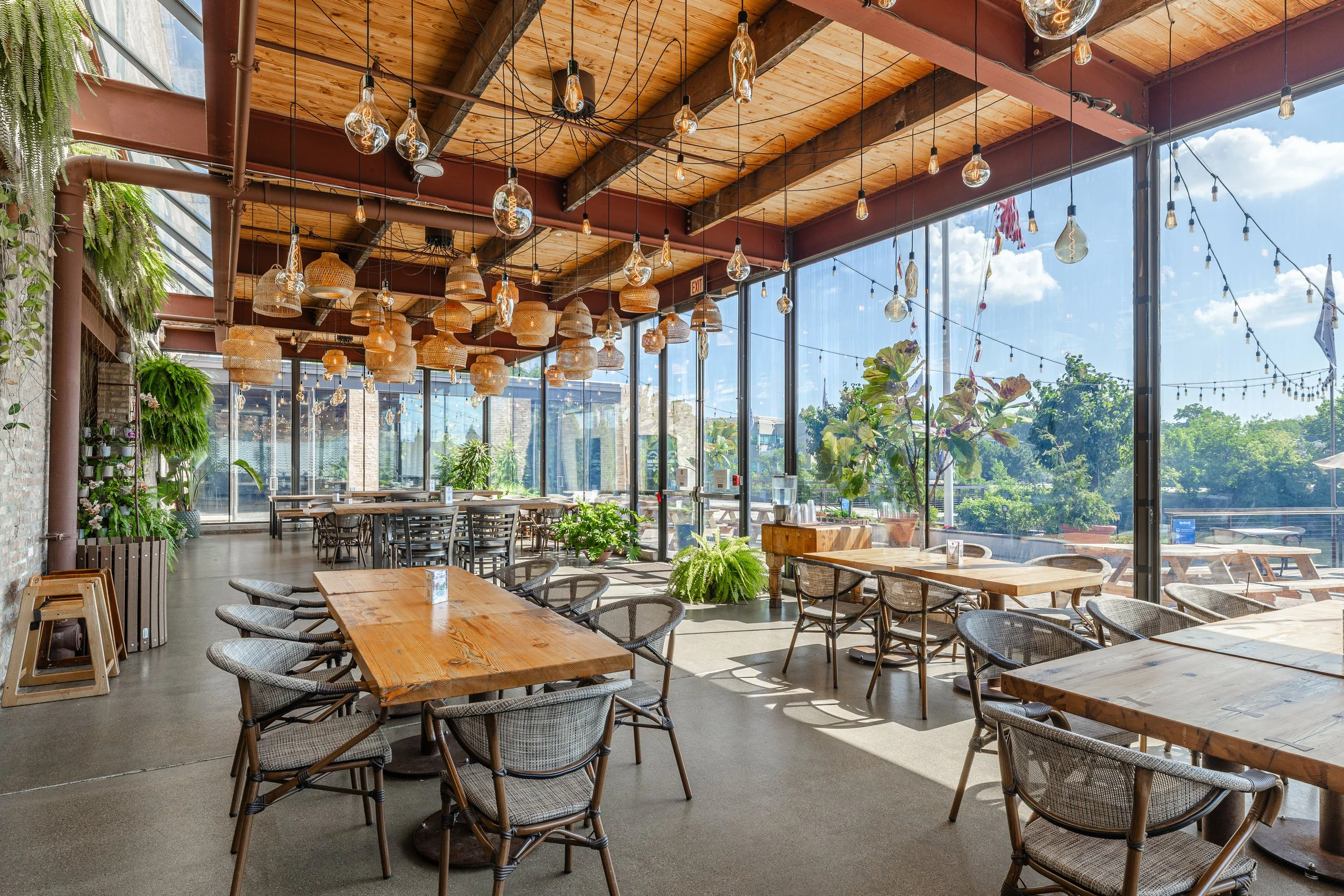 Bright indoor restaurant space with wooden tables, wicker chairs, potted plants, and large windows with outdoor seating visible outside.