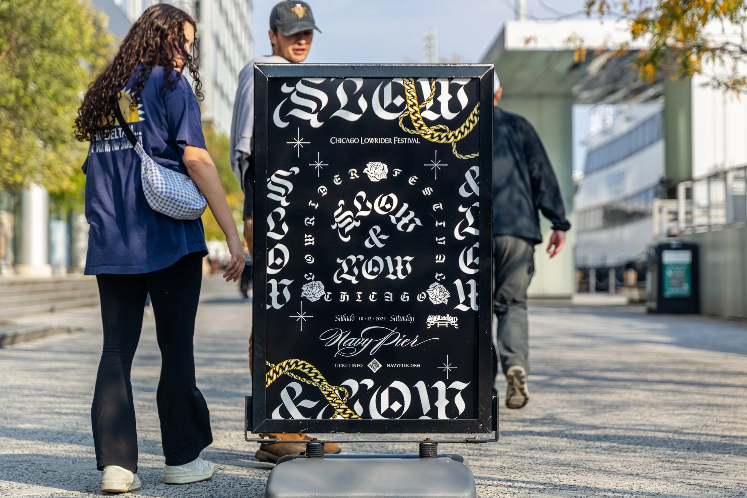 A black signboard on a sidewalk advertising the Chicago Lowrider Festival, including details about the event date, location, and website, with design elements like gold chains and roses, and people walking past in the background.