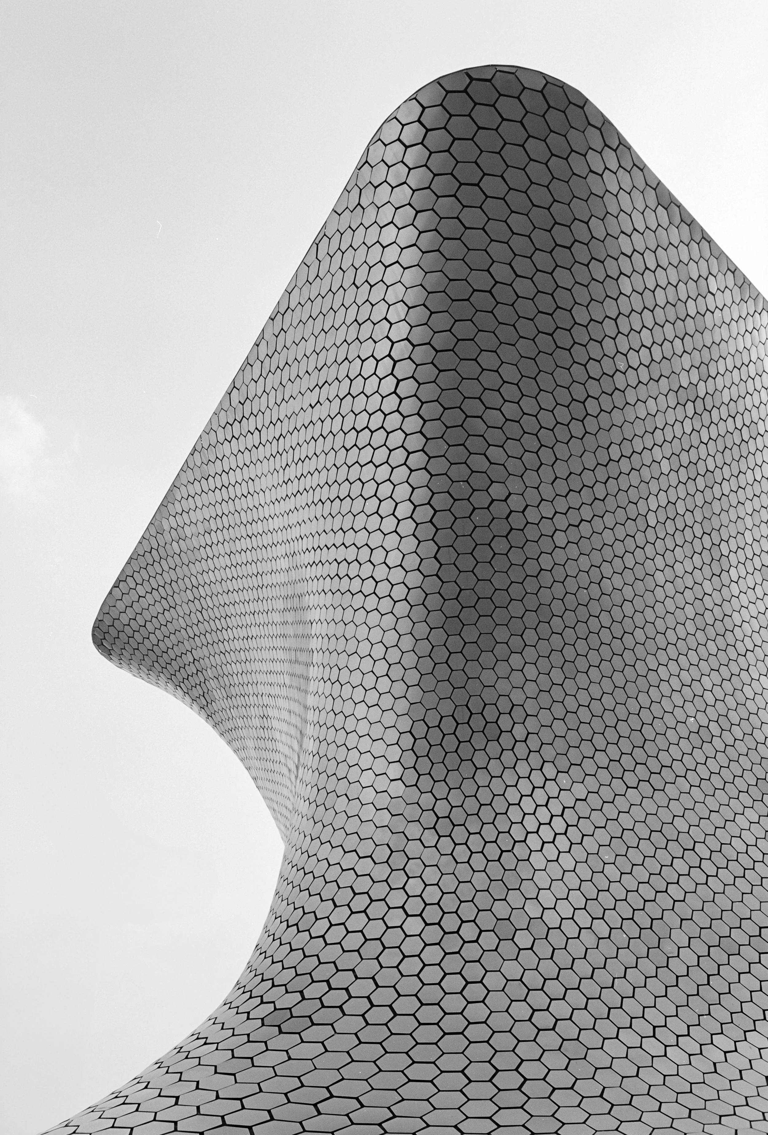 Close-up view of the uniquely curved exterior of a modern building covered in hexagonal tiles, against a light sky background.