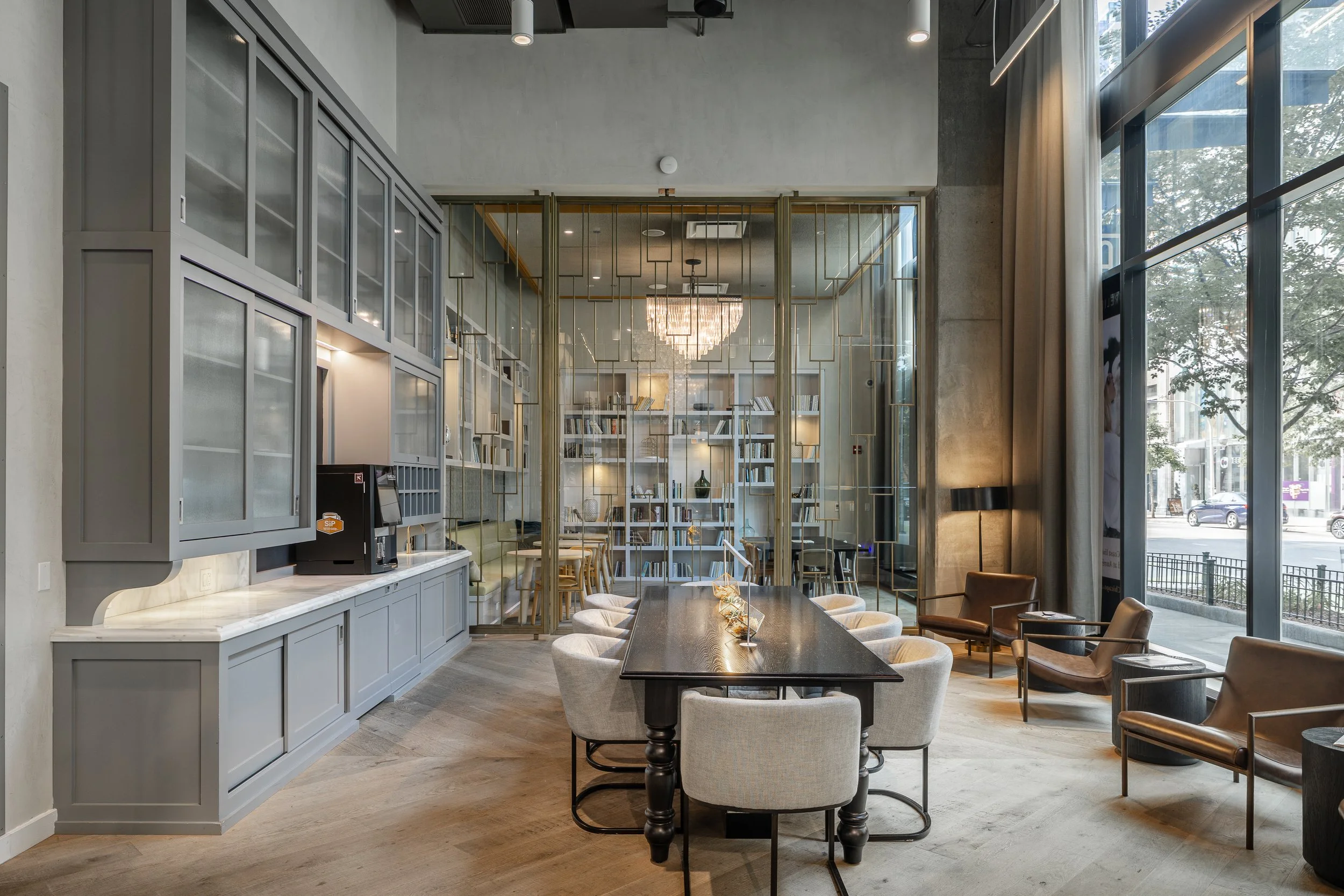 Modern hotel lobby with a large central table surrounded by cream-colored chairs, a bookshelf behind a glass partition, a chandelier hanging from the ceiling, and large windows on the right side letting in natural light.