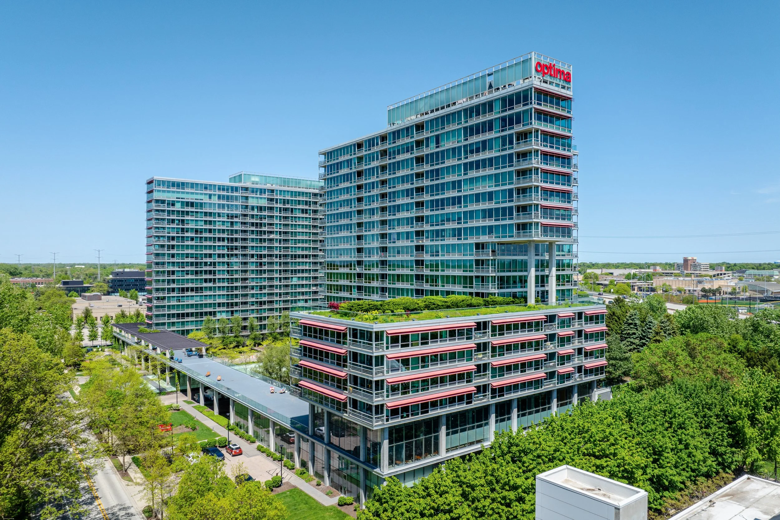 Modern multi-story glass office buildings with greenery and parking lot in foreground, clear blue sky in background.
