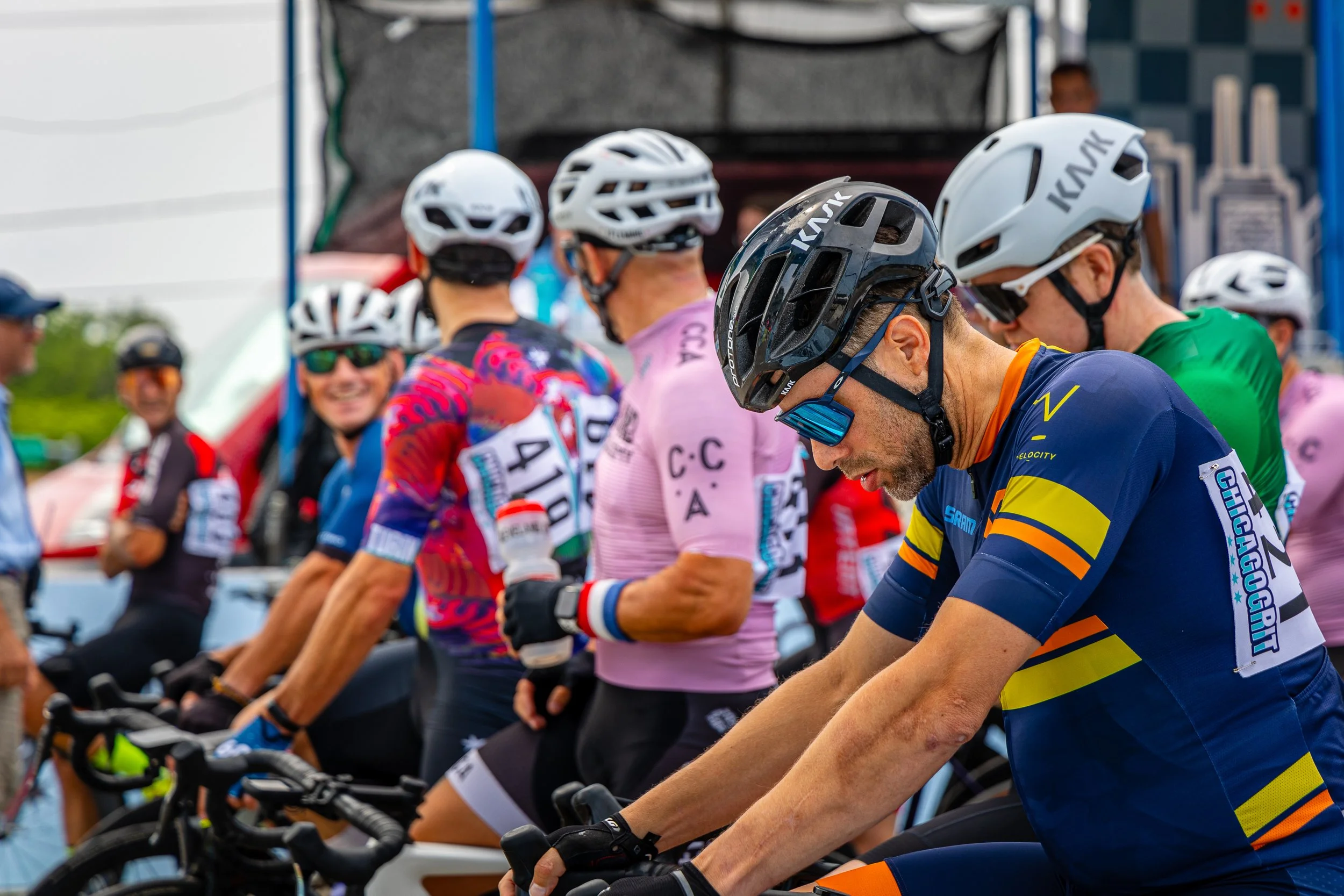Cyclists dressed in colorful jerseys and helmets preparing for a race, standing with their bikes at the starting line.