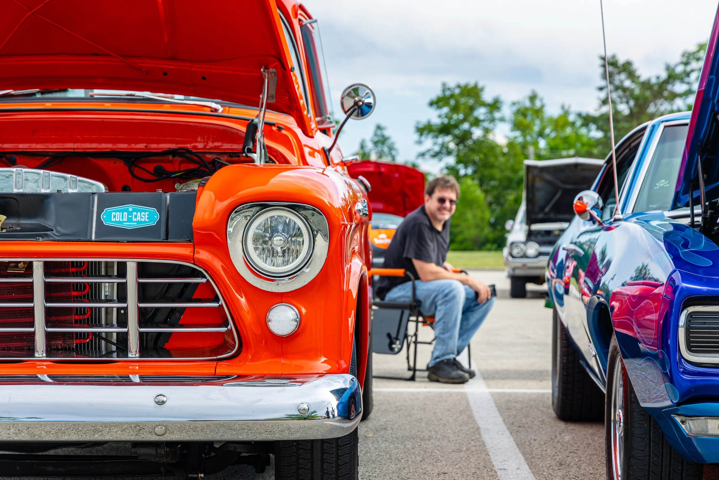 Orange vintage pickup truck with its hood open parked at a car show, with a man sitting on a chair nearby and other classic cars in the background.
