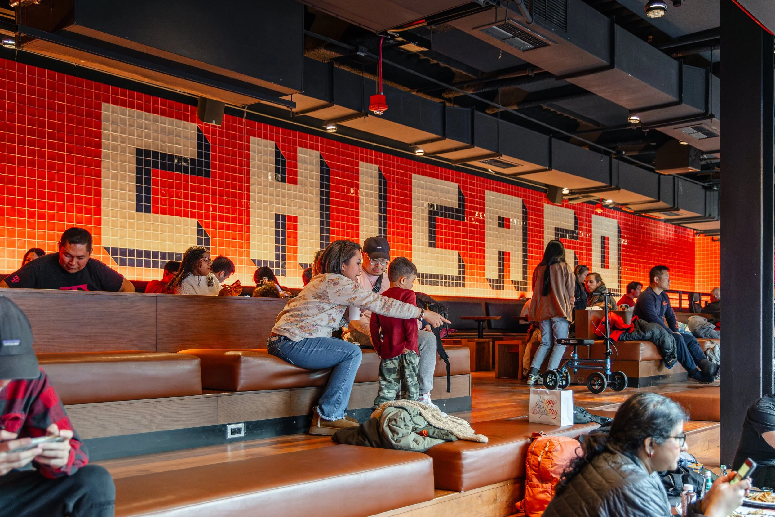 People sitting and standing inside a modern restaurant or cafe, with a large red mosaic wall spelling out 'SILVERADO', and various seating arrangements.