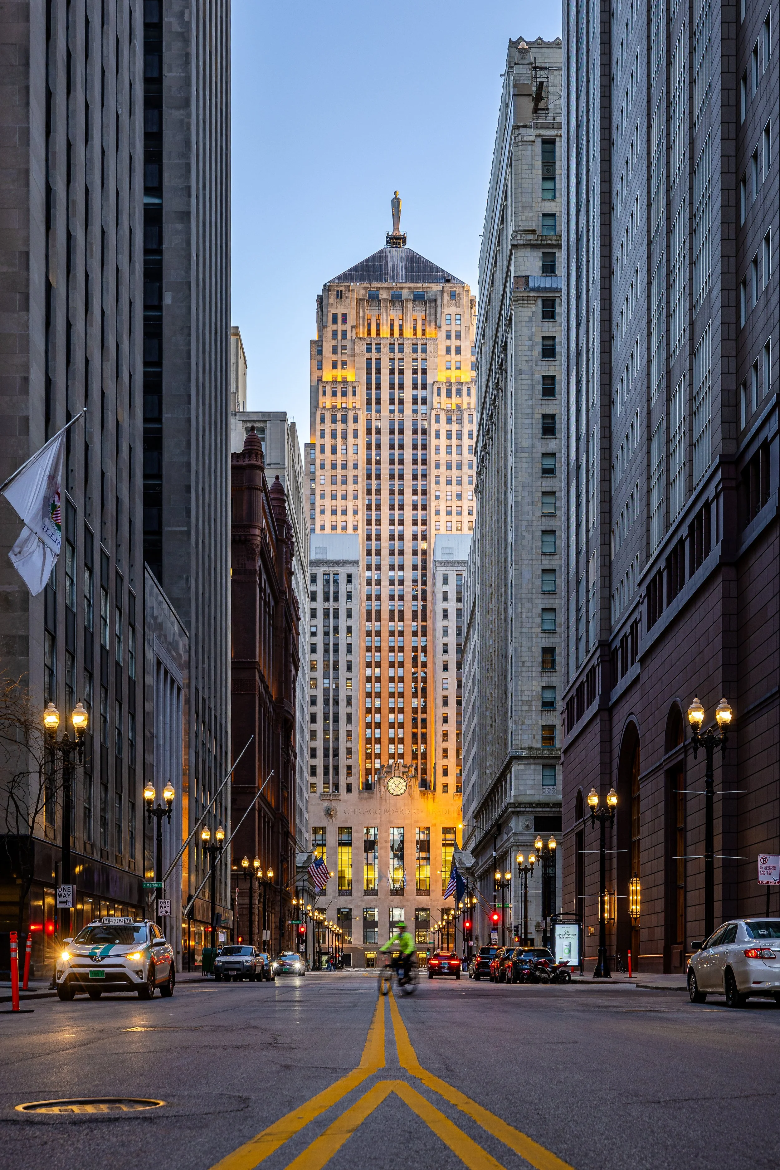 A city street at dusk with the Chicago Board of Trade Building illuminated in the background, cars parked and driving, a cyclist in the middle, and street lamps lining the sidewalk.