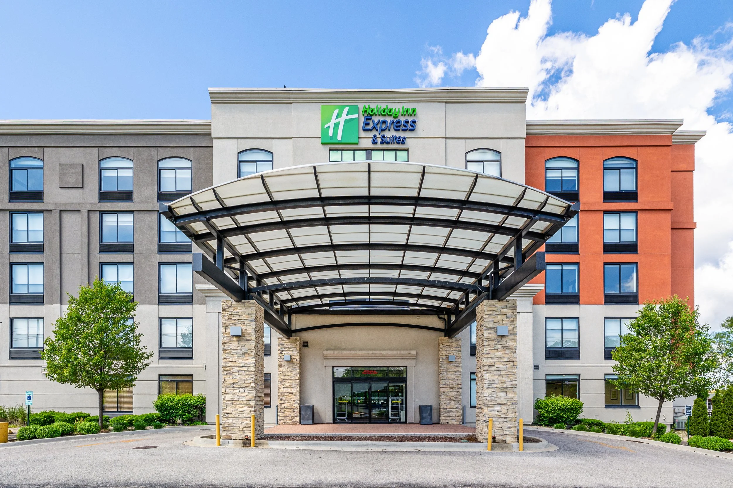 Front entrance of a Holiday Inn Express & Suites hotel with a covered drop-off area, trees, and bushes, under a partly cloudy blue sky.