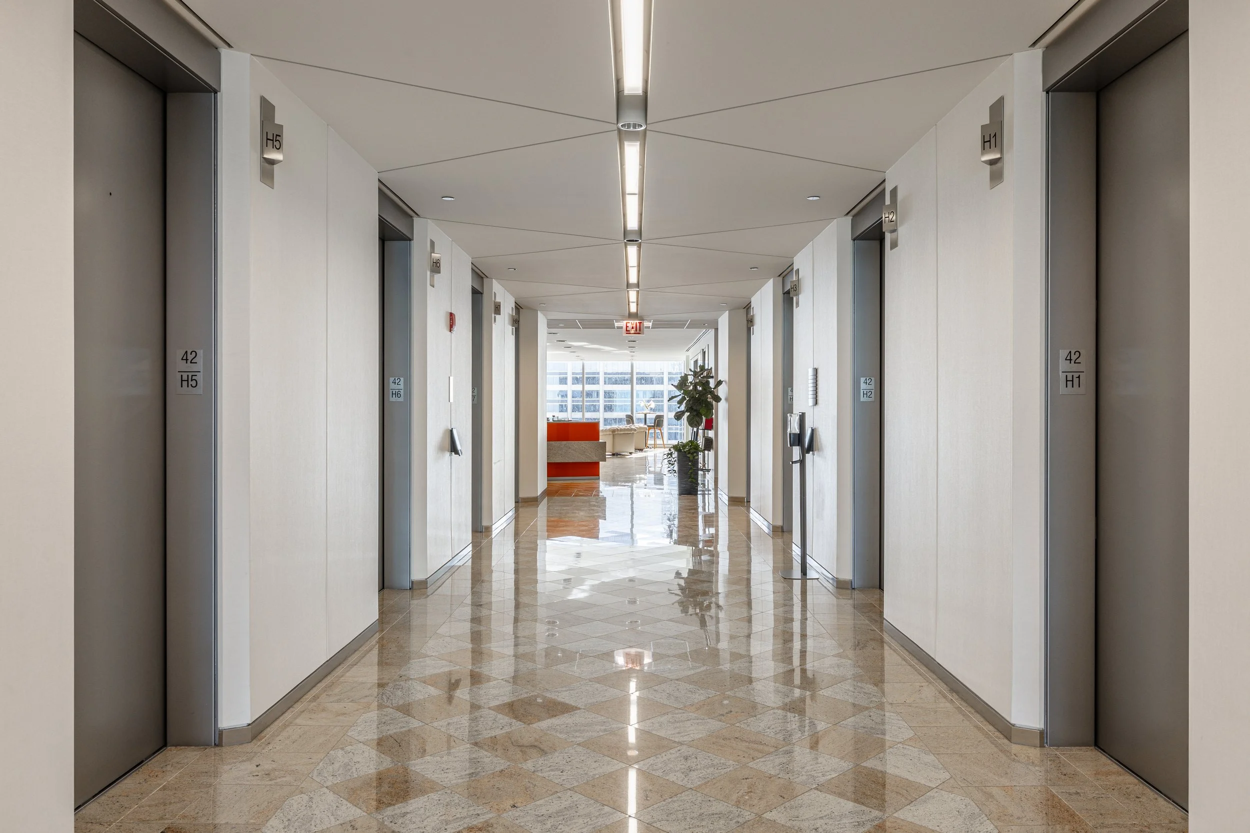 Interior view of a modern elevator lobby with multiple elevator doors on both sides, numbered H1 to H6, and a seating area with chairs and a table in the background.