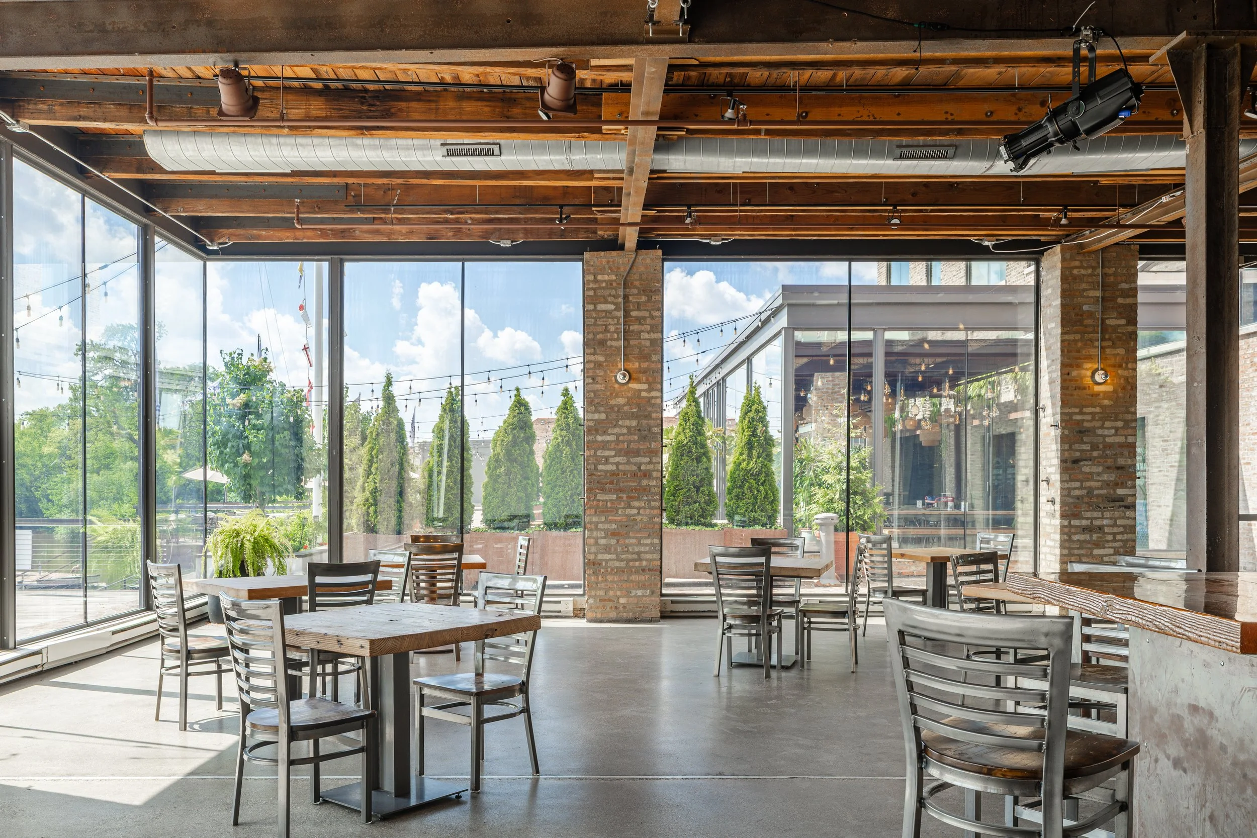 Empty restaurant interior with large glass windows, wooden ceiling, brick pillars, and metal chairs around wooden tables, with outdoor greenery visible outside.
