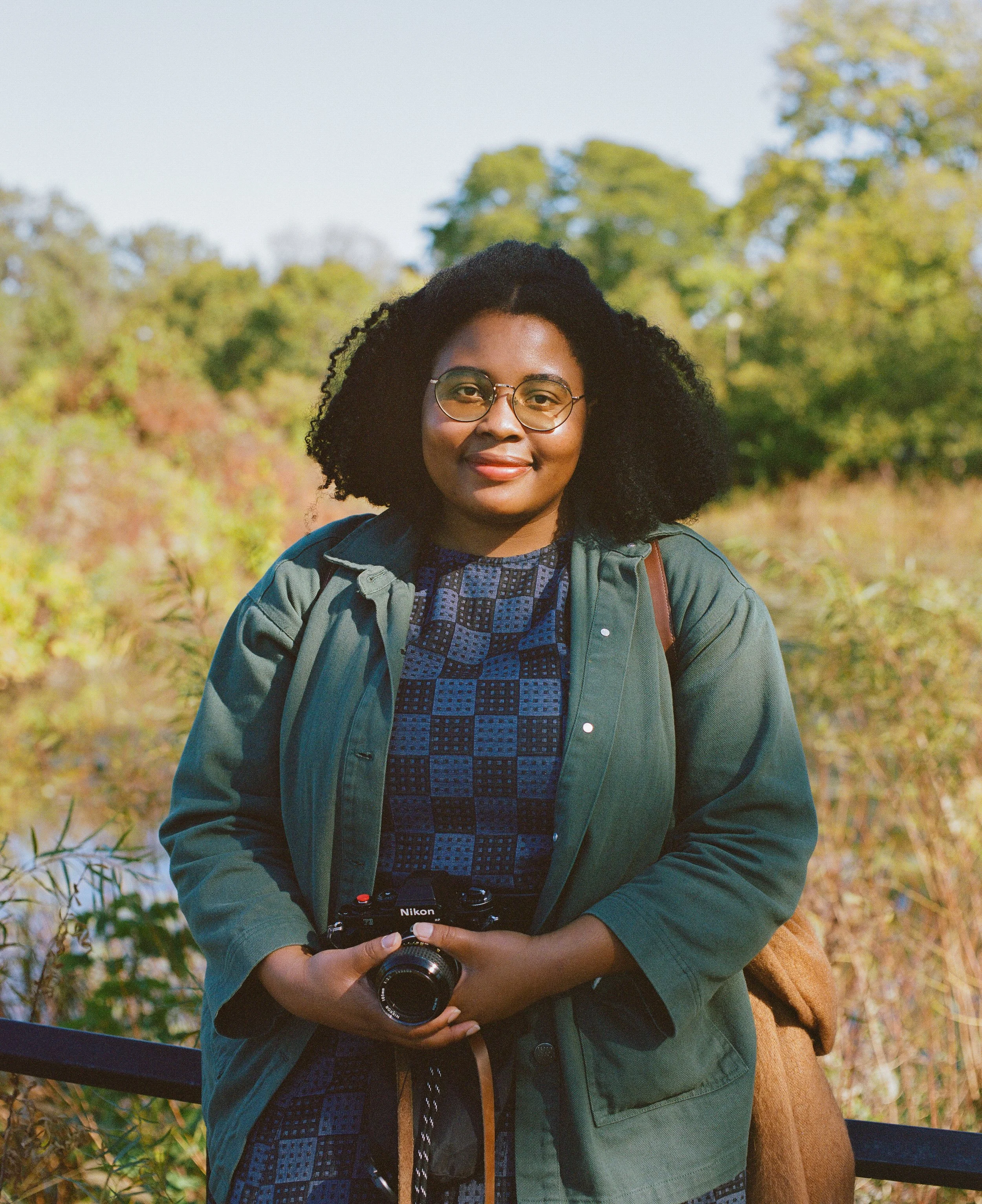 A woman outdoors with trees and foliage in the background, holding a Nikon camera, wearing glasses, a patterned dress, and a green jacket.