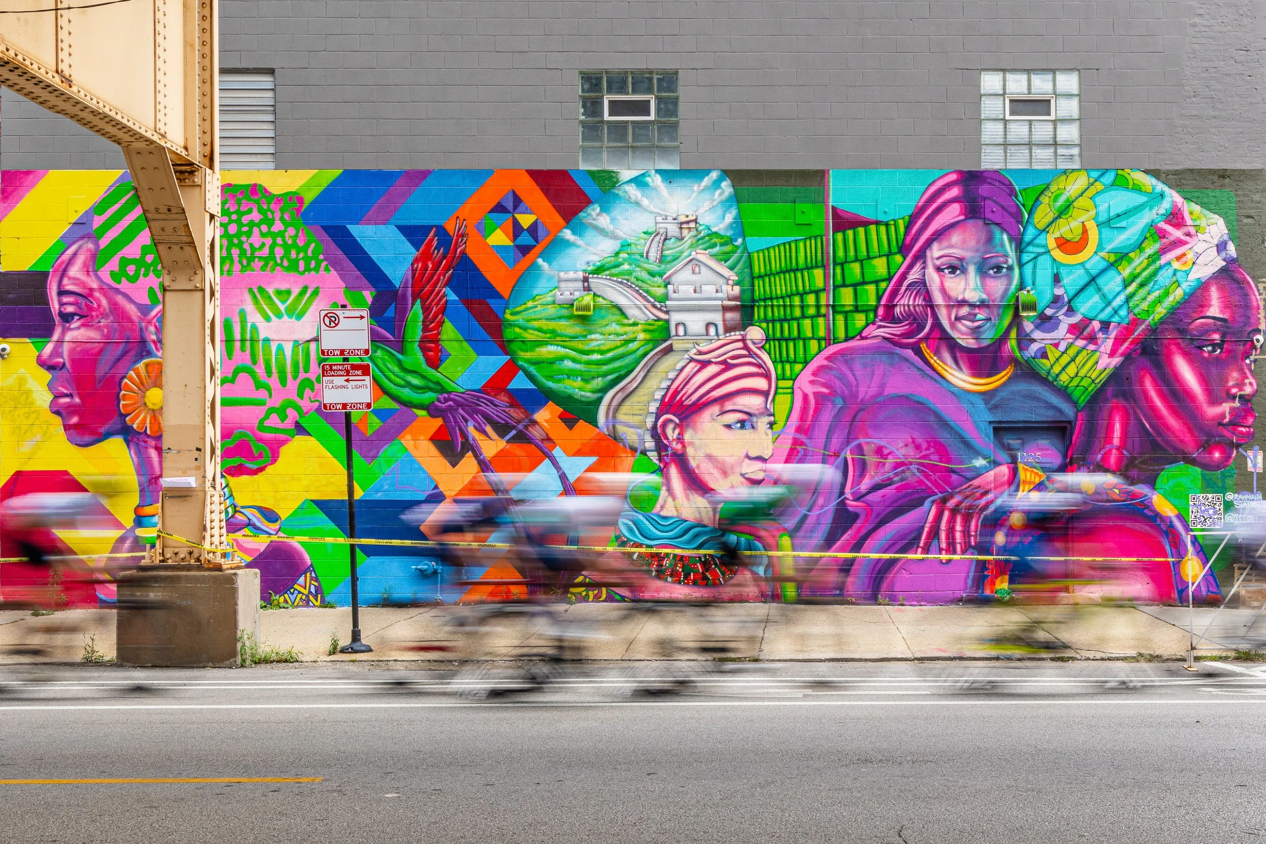 Colorful mural featuring diverse women with vibrant backgrounds and cultural symbols, painted on a city wall along a street with moving traffic.