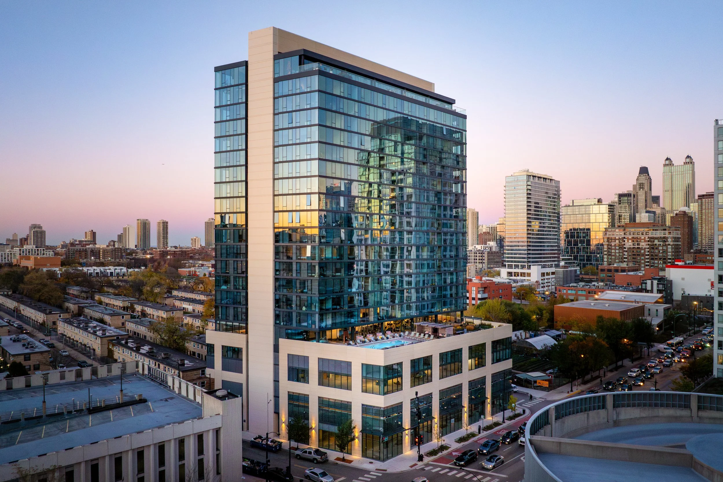Modern glass high-rise building in a cityscape during sunset, with reflections of other buildings on its windows and surrounding urban environment.