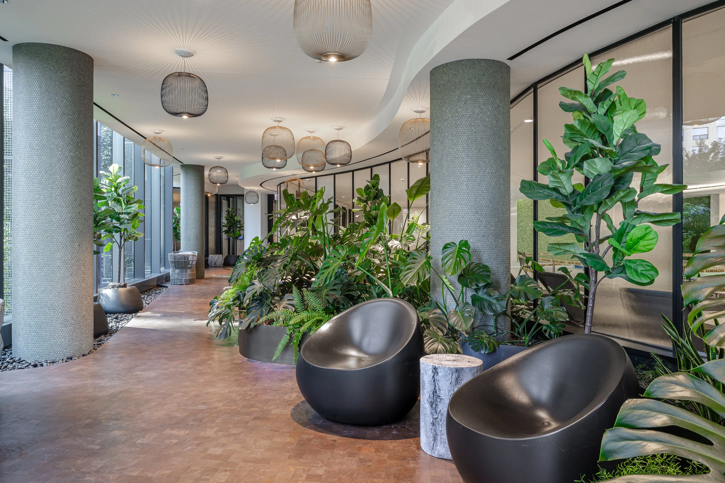 Modern indoor space with large green plants, black lounge chairs, and hanging ceiling lights.