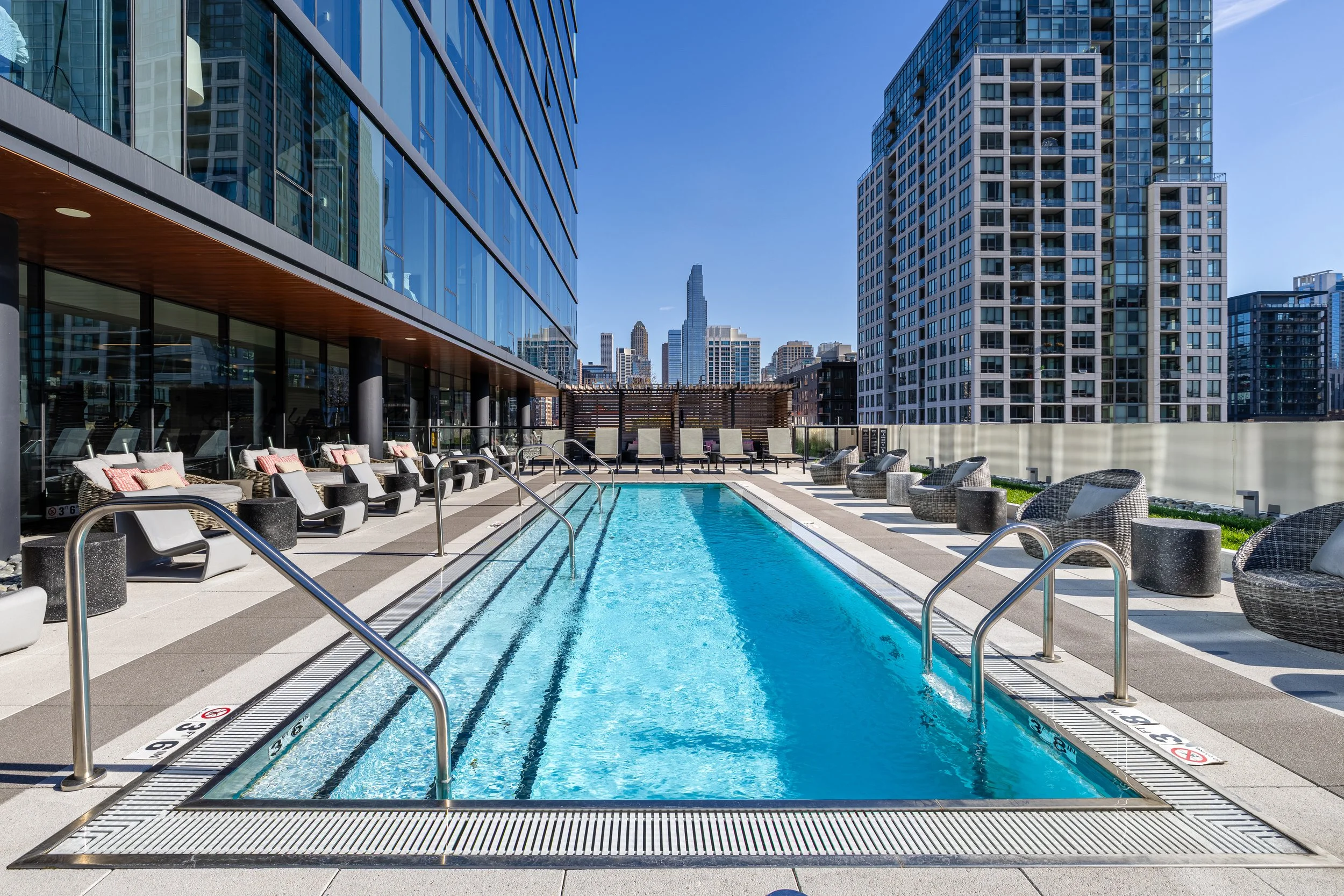 Rooftop pool area with lounge chairs and wicker seating surrounded by tall city buildings under a clear blue sky.