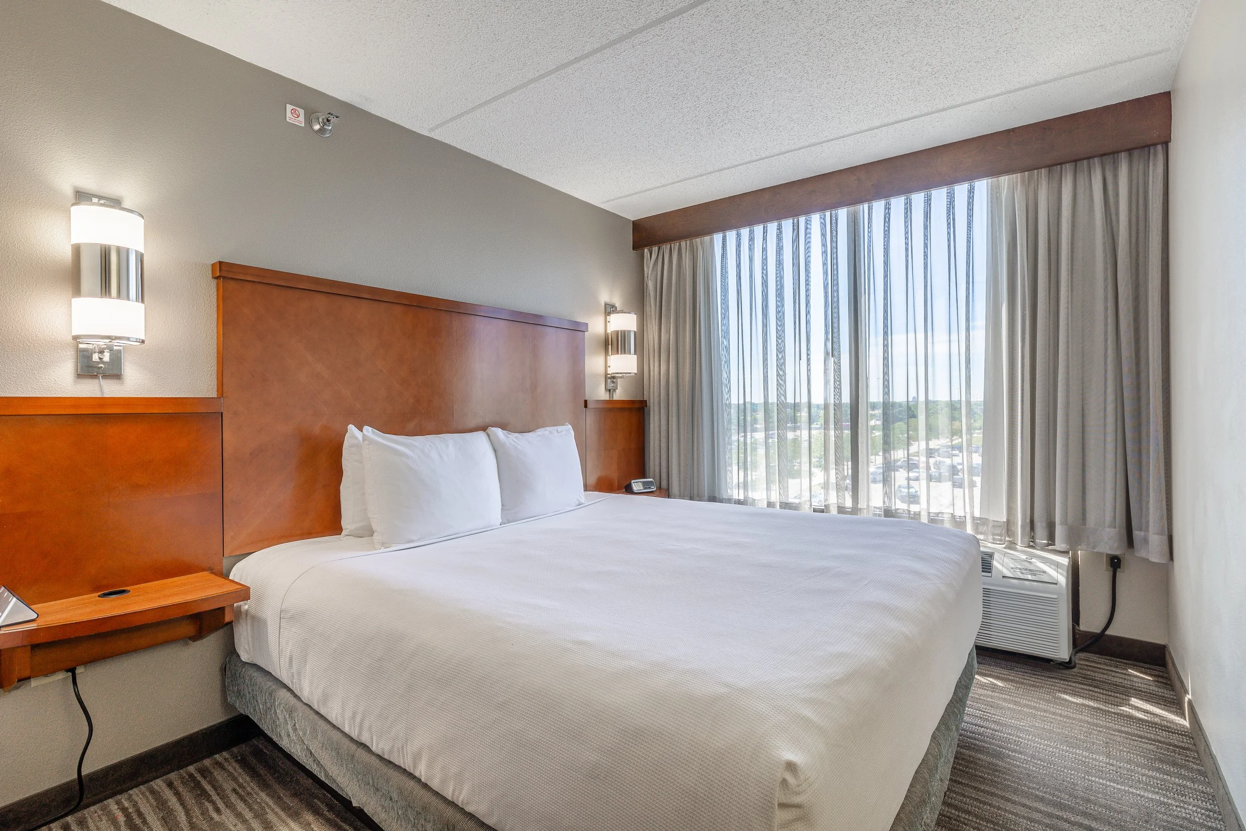 A hotel room with a neatly made bed, white linens, two pillows, wooden headboard, wall-mounted lights, a small nightstand with a phone, large window with sheer curtains revealing a view of parking lot and trees, and a window air conditioning unit.