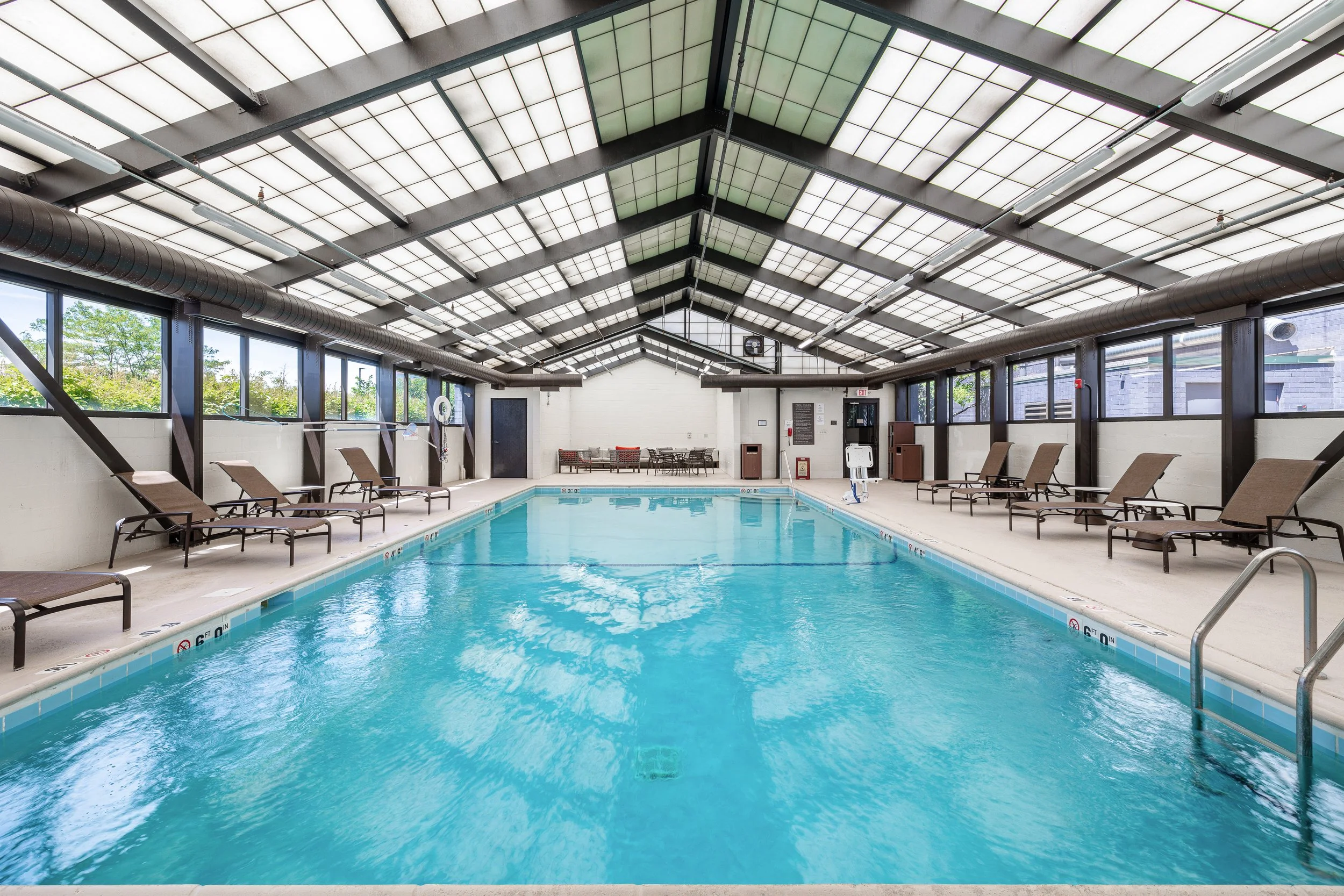 Indoor swimming pool area with lounge chairs along the sides, a covered roof, and windows allowing natural light, with a seating area at the far end.