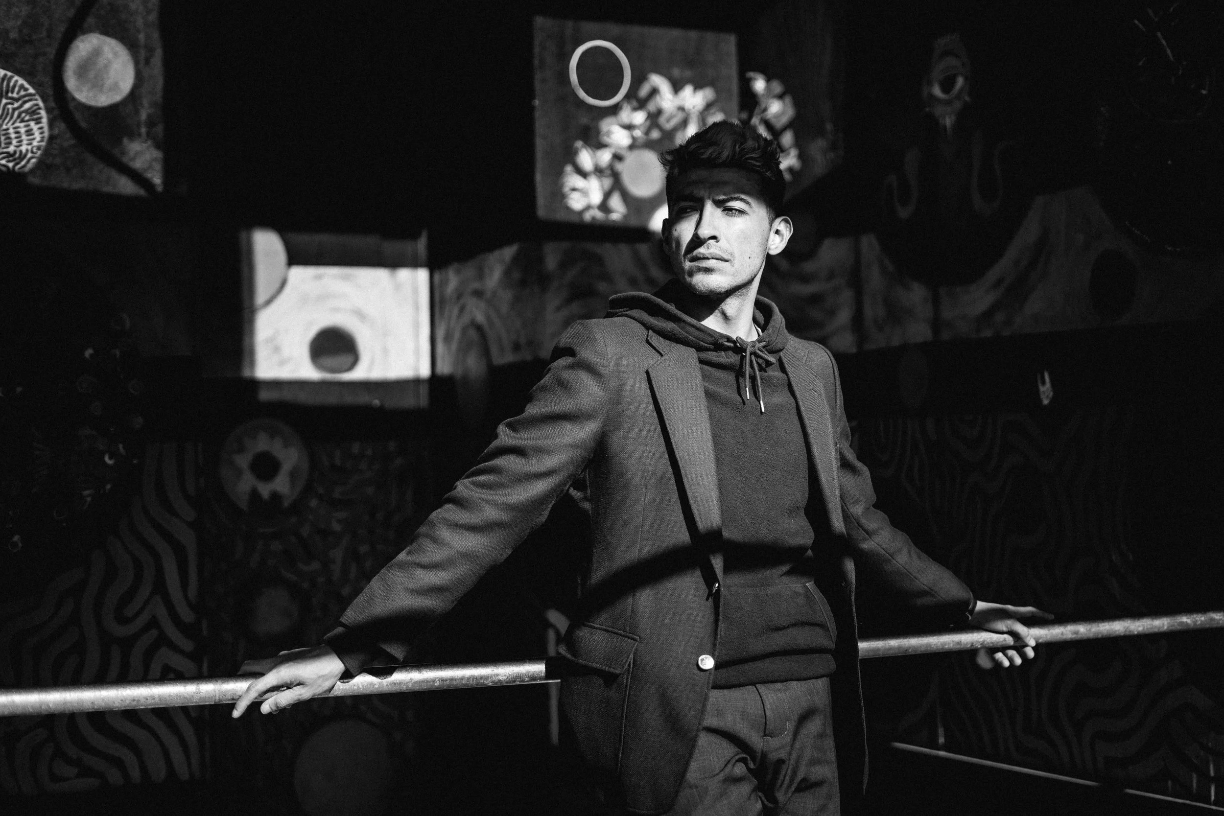 A young man in a blazer and hoodie stands in a dark space, leaning on a metal railing with graffiti art in the background, looking to the side.