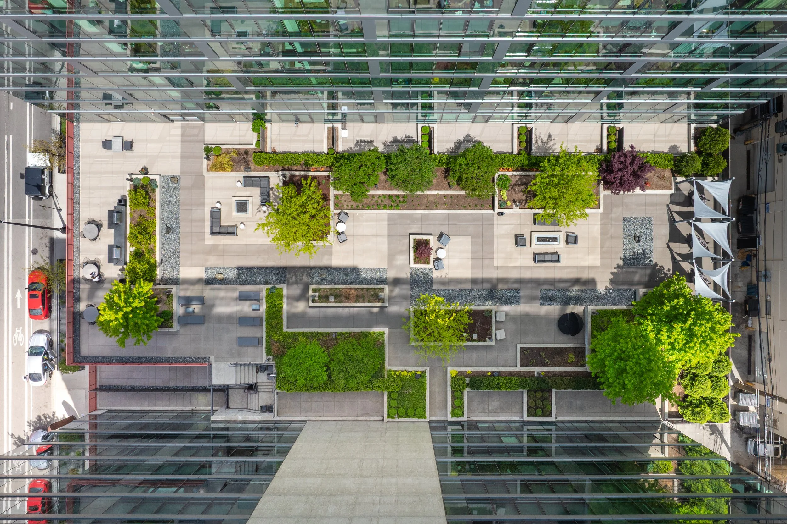 Aerial view of an urban rooftop garden with trees, plants, outdoor seating, and shaded areas surrounded by glass buildings and city streets.
