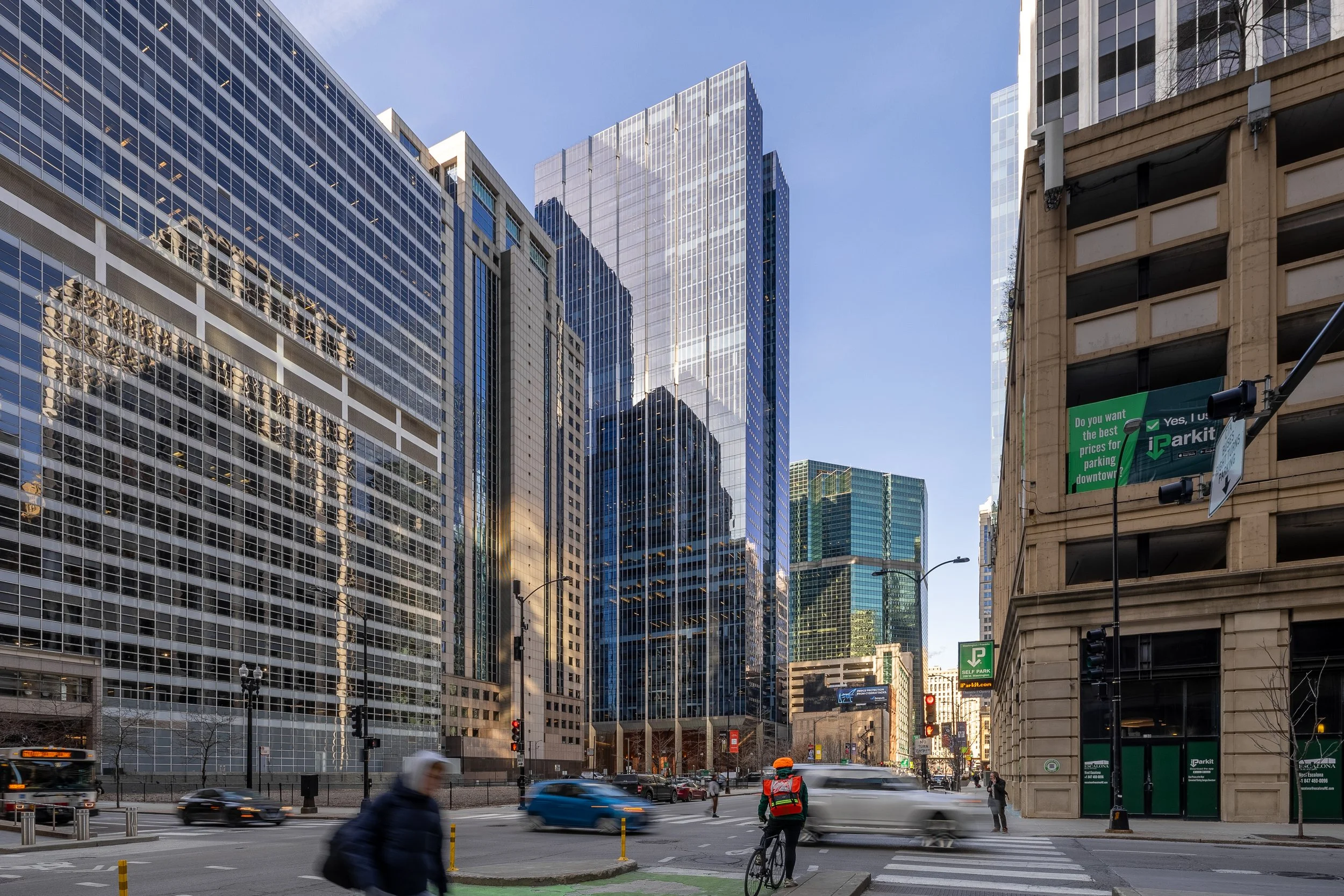 City street with tall glass skyscrapers, a cyclist in an orange helmet, pedestrians crossing, cars, and a parking garage on the right side.