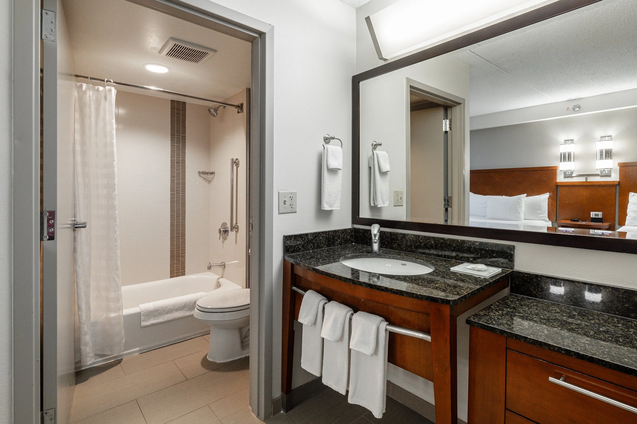 View of a hotel bathroom with a vanity, large mirror, handheld towel, and a toilet and bathtub in the background.