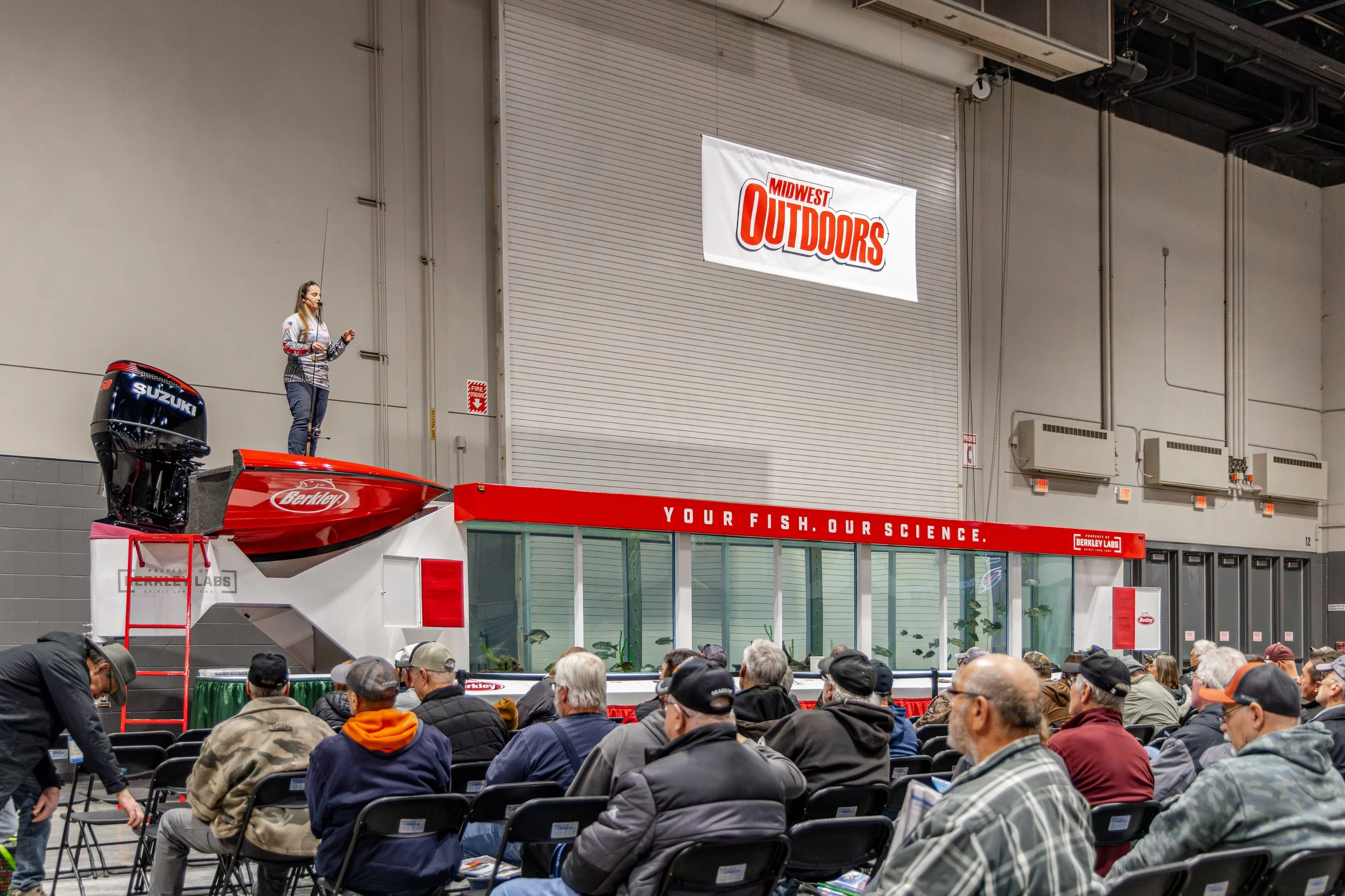 A speaker standing on a red platform in front of an audience at an indoor event, with banners displaying 'Midwest Outdoors' and 'Your Fish. Our Science.' The speaker is next to a fish tank exhibit with fish inside.