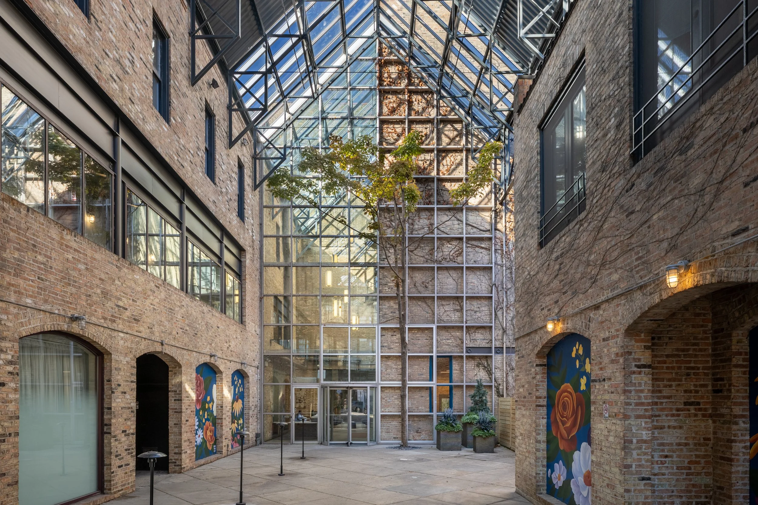 An indoor courtyard with brick walls, large glass windows and a glass roof. There is a tall tree in the center, and colorful mural paintings on the walls.