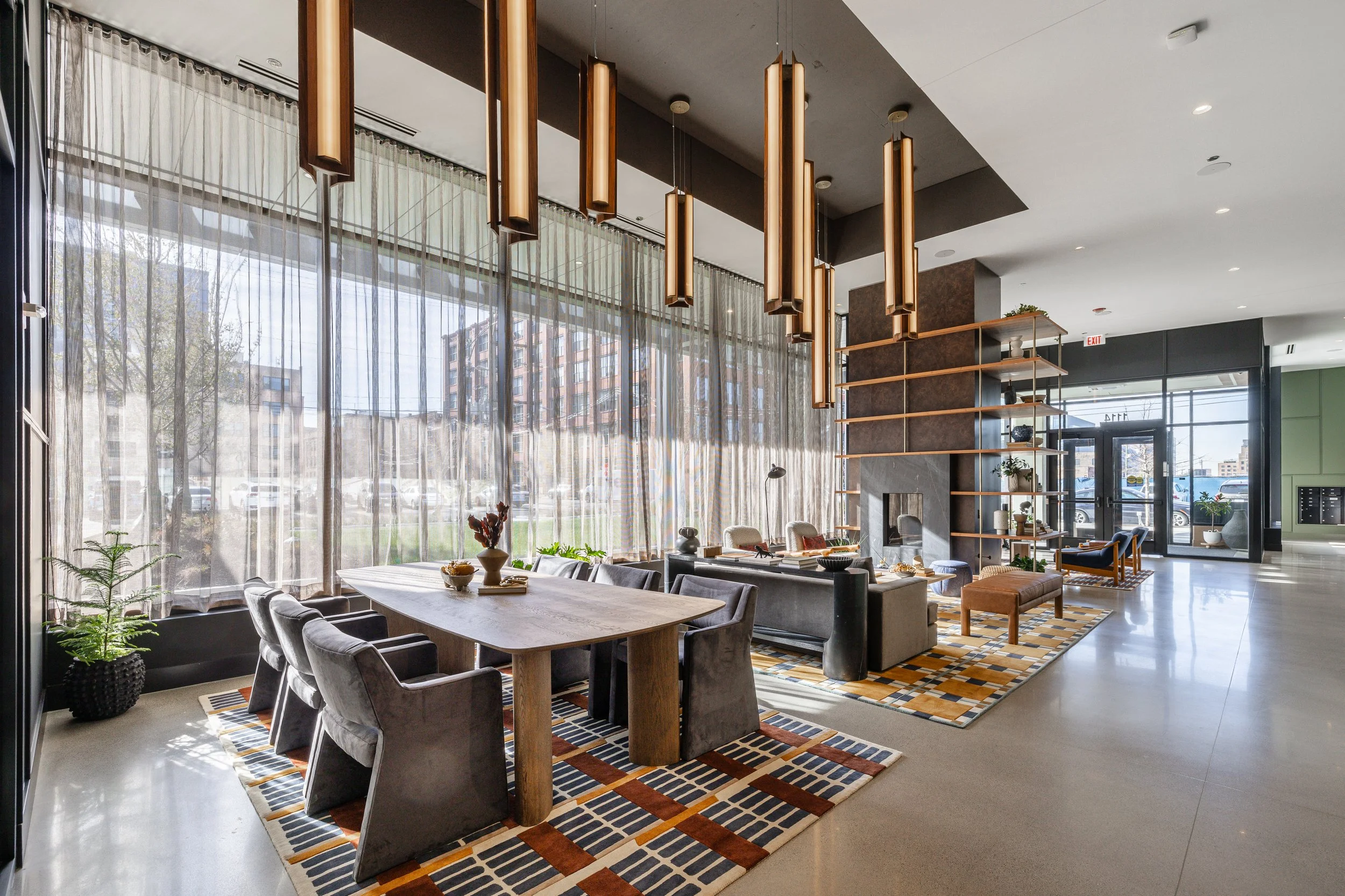 Interior of a modern lobby with a dining area, large windows with sheer curtains, contemporary lighting, and a seating area with sofas, chairs, and decorative shelves.