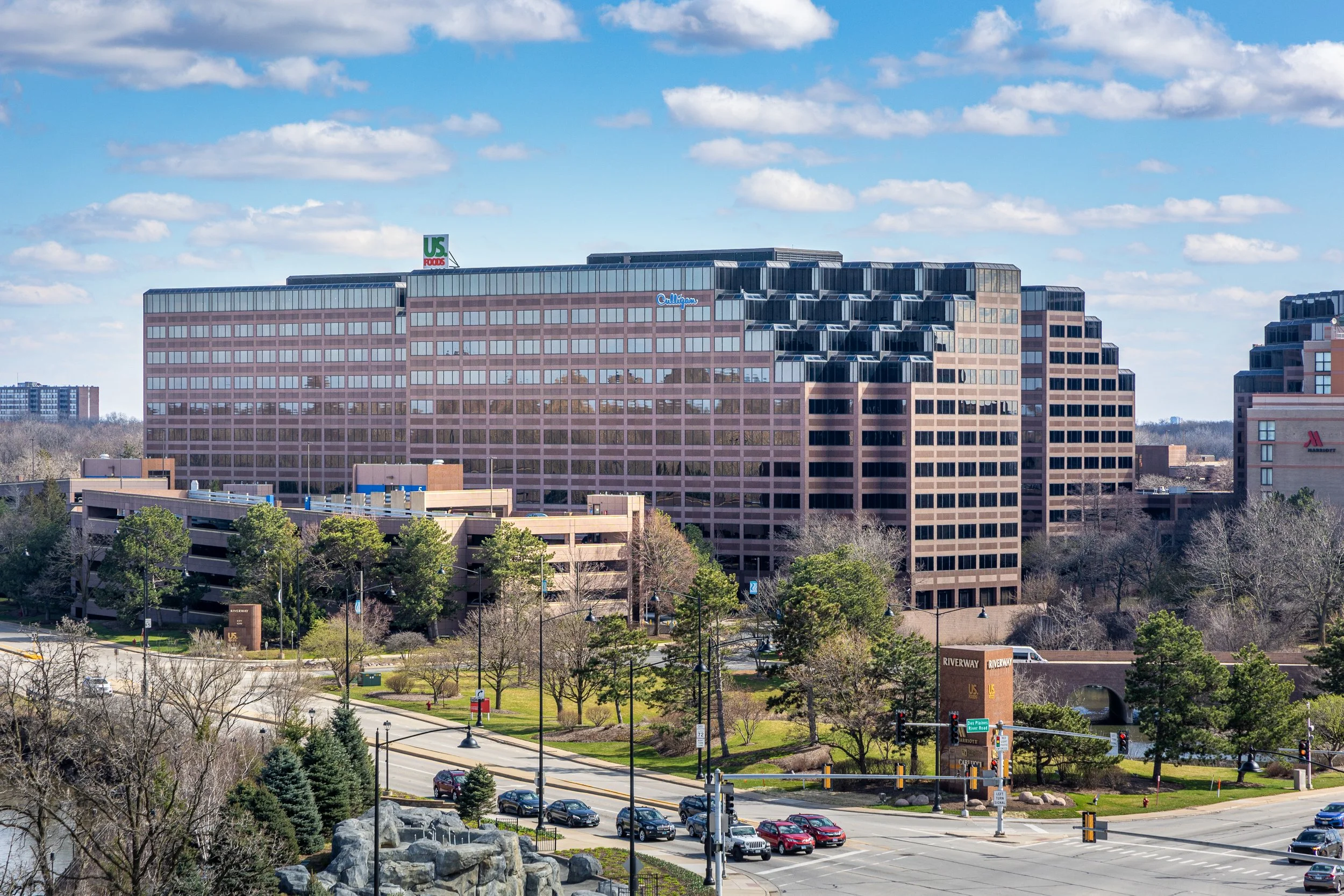 A large office building with multiple floors, glass windows, and signage including U.S. Foods and Milliken. Surrounding the building are trees, a parking garage, and a street with cars.