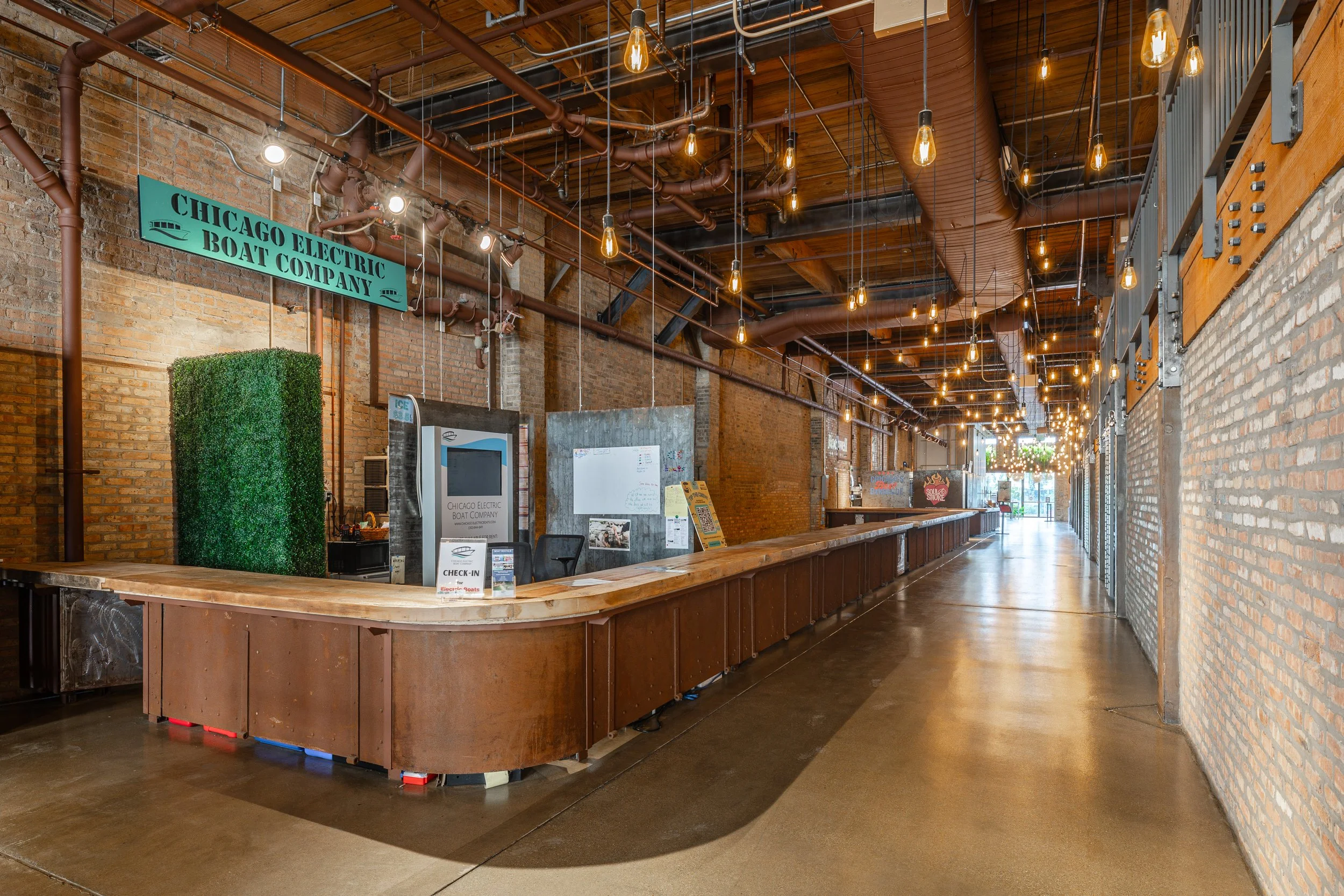 Empty reception area with a long wooden counter, exposed brick walls, hanging Edison bulb lights, and industrial piping on the ceiling.