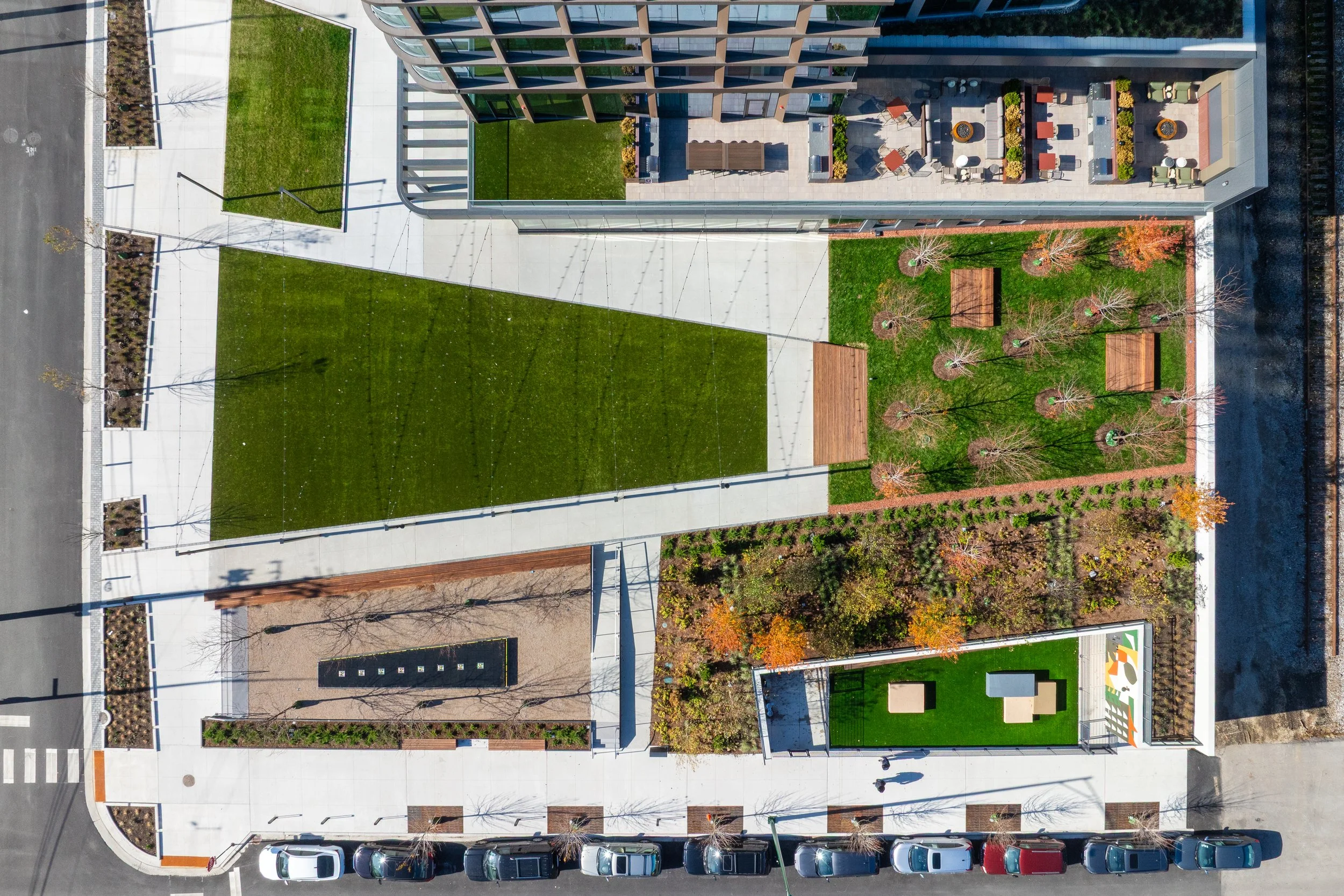 Top-down aerial view of a modern urban park with grassy areas, trees, benches, and surrounding paved walkways. Adjacent building features a rooftop terrace with seating and tables.
