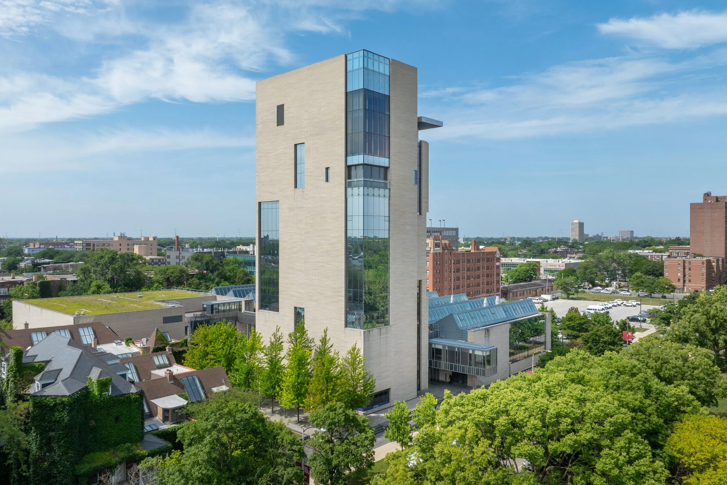 Modern multi-story building with glass windows and light-colored facade surrounded by trees in an urban area under a blue sky with scattered clouds.