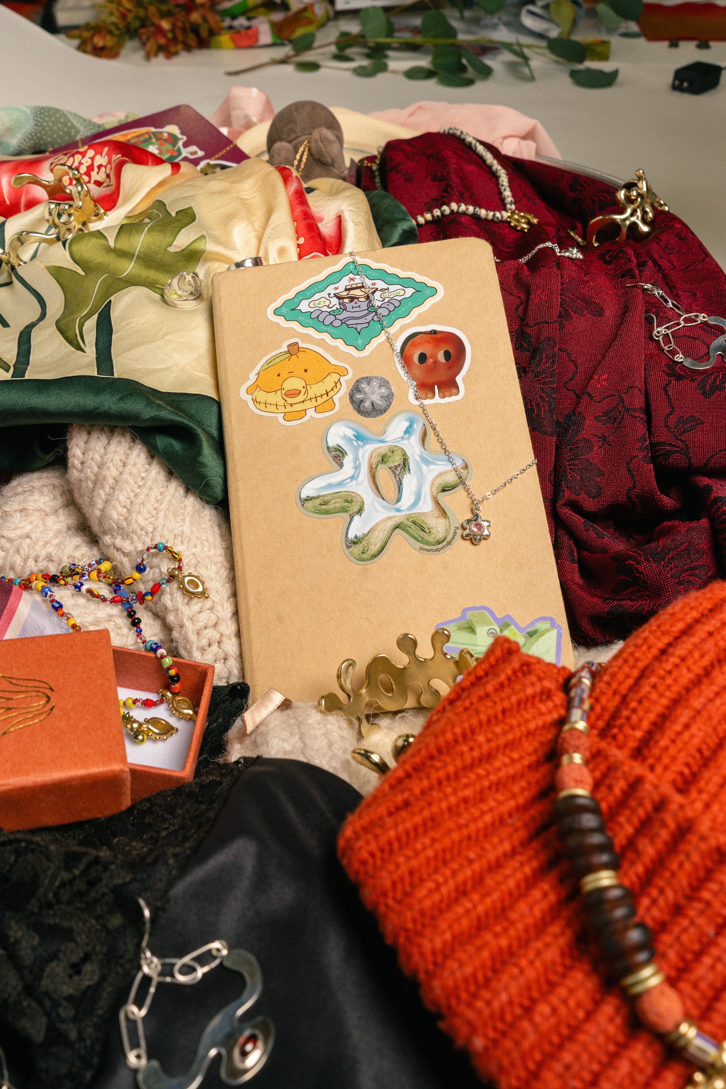 Various jewelry and accessories scattered around a cardboard display board with stickers and necklaces, including a red and black fabric, knitted gloves, and a decorative orange cloth.