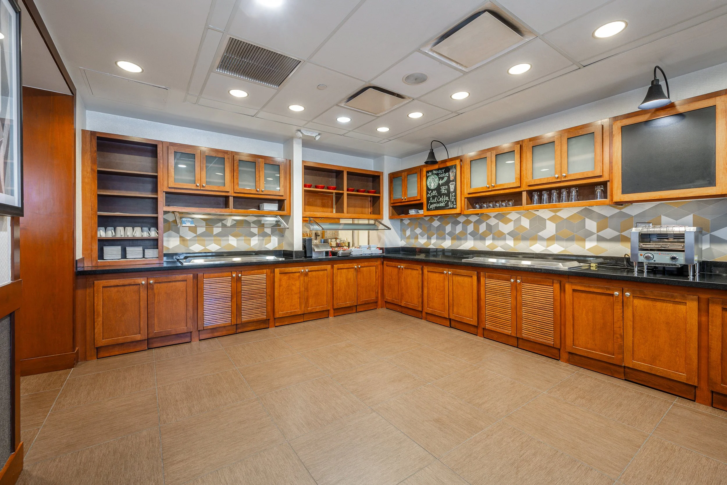 Empty commercial kitchen with wooden cabinets, black countertops, geometric patterned backsplash, and beige tiled floor.