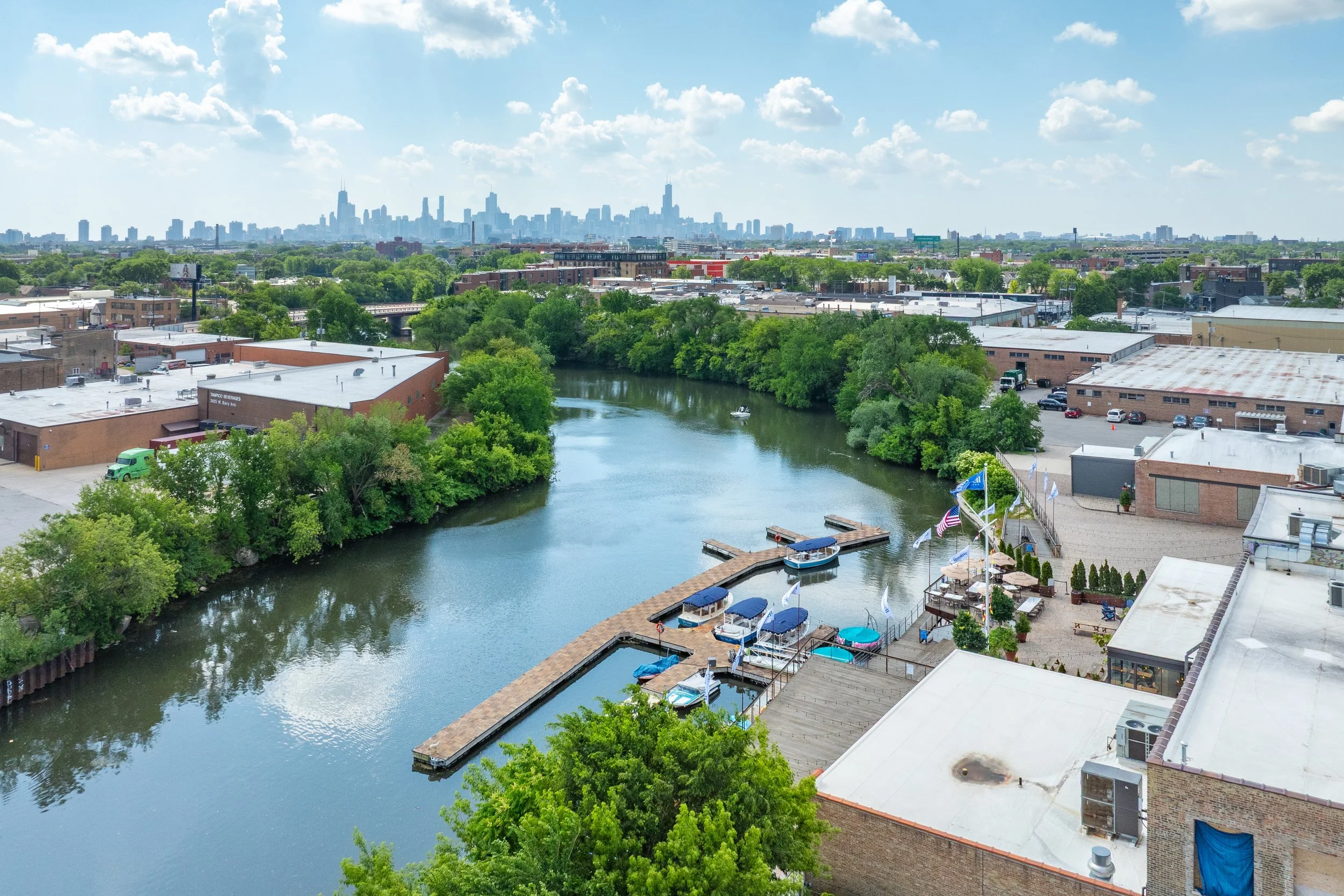 A river with trees on its banks runs through an urban area with a mix of industrial and commercial buildings, a boat docked at a marina, and a city skyline in the background under a partly cloudy sky.