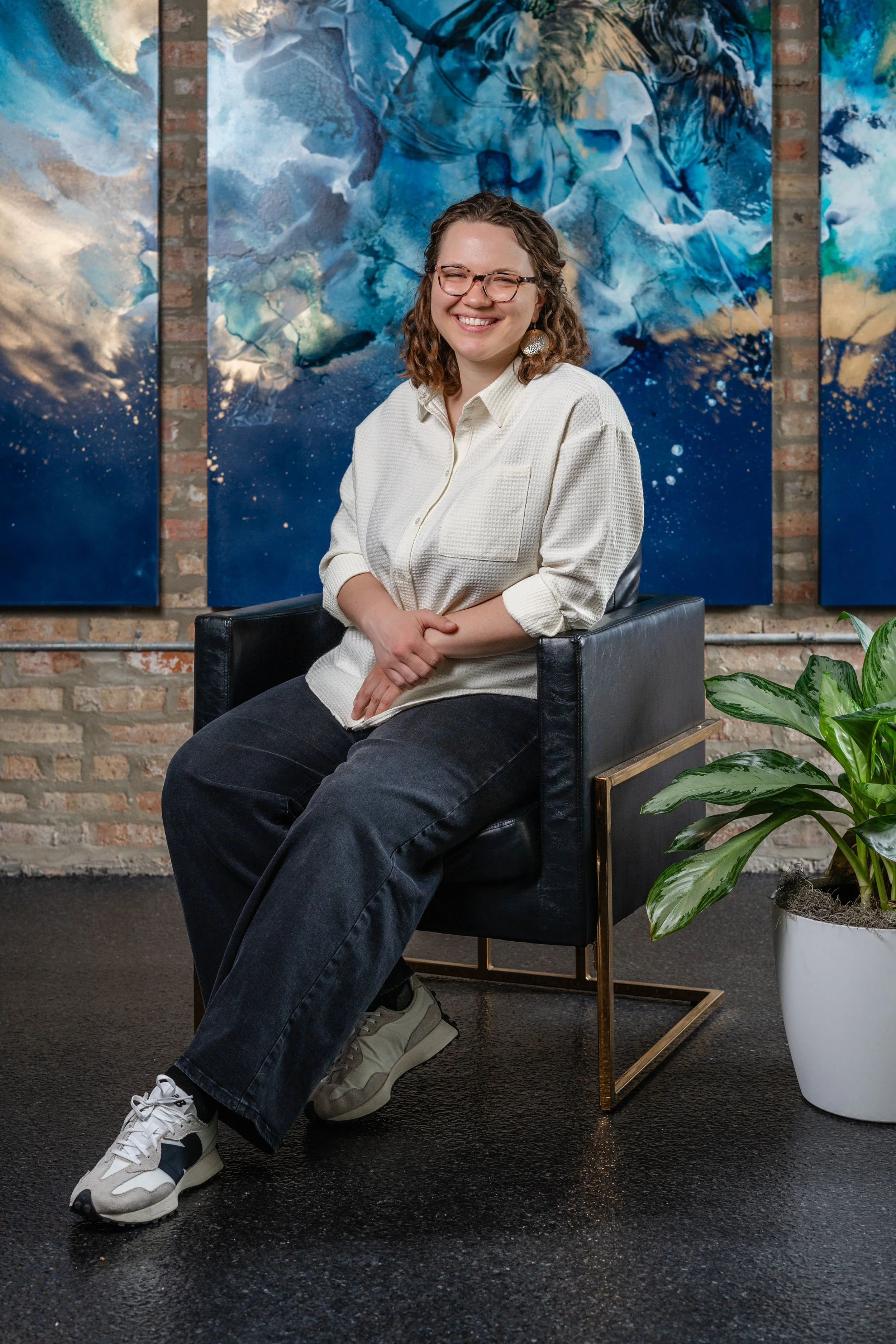 A woman with curly hair, wearing glasses, a white button-up shirt, and black pants, sits on a black armchair smiling at the camera. Behind her are abstract blue and white paintings on a brick wall. To her right is a green leafy plant in a white pot.