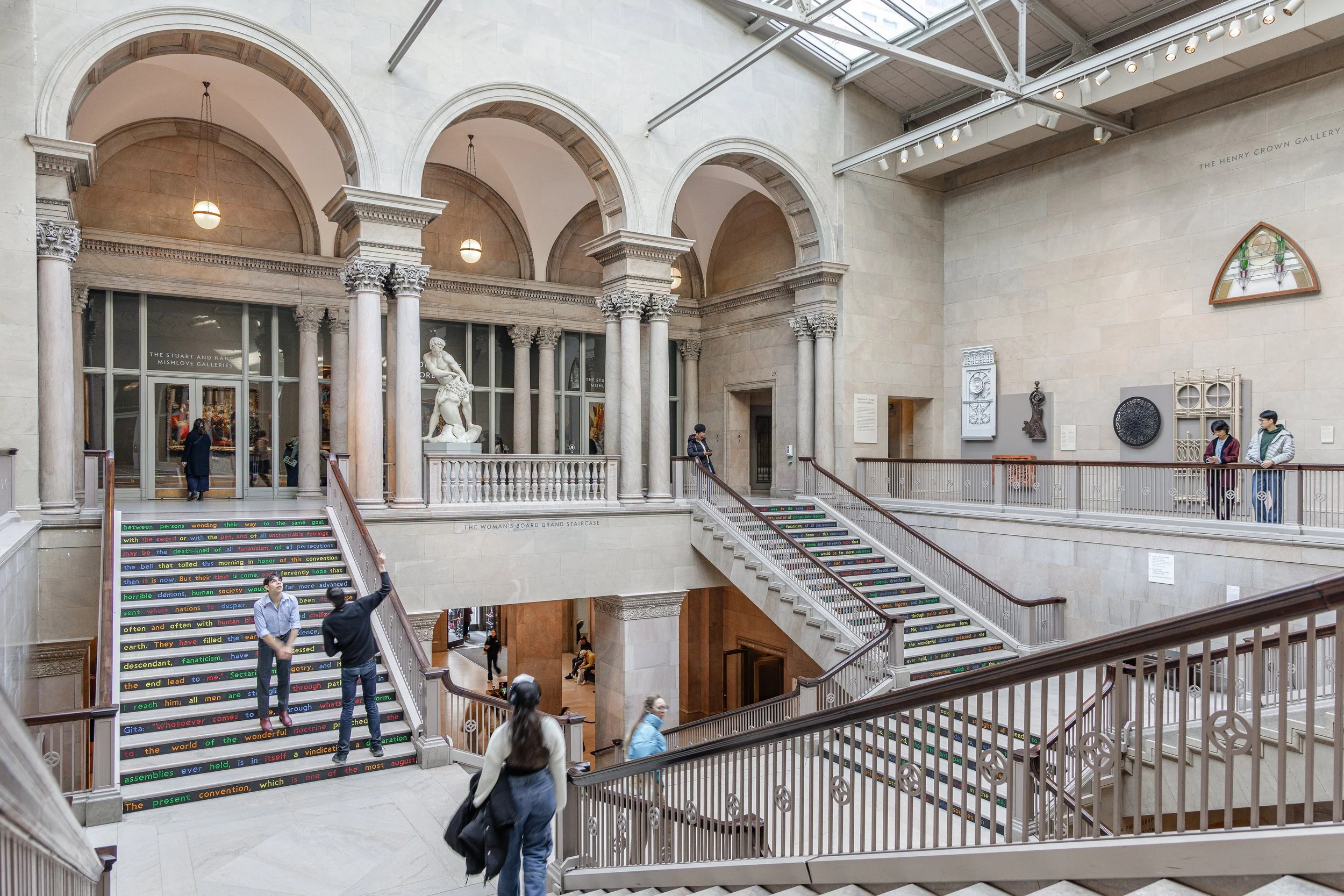 Interior of a museum with grand staircase, people walking, viewed from the second floor looking down.