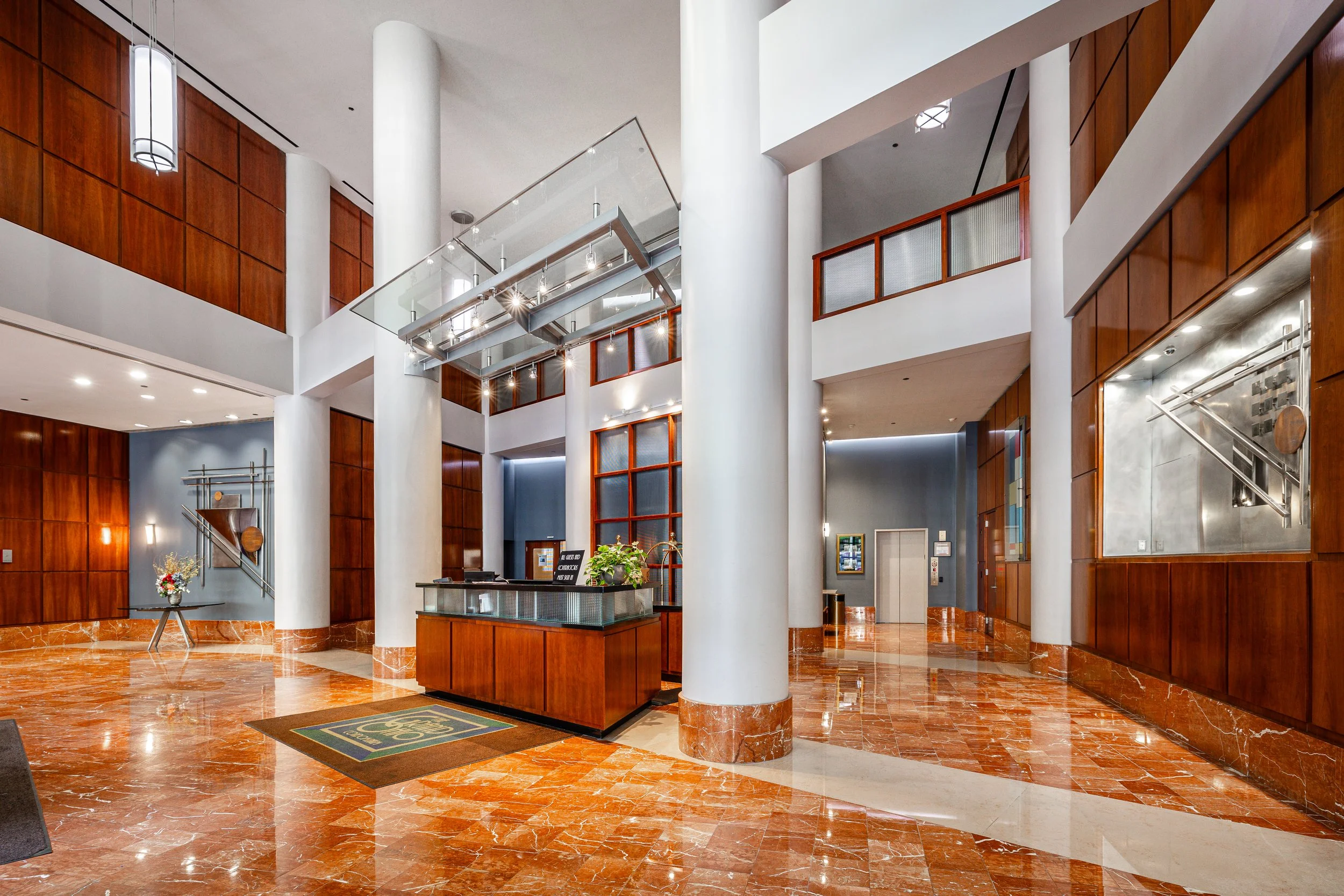 Hotel lobby with marble floors, wooden walls, a reception desk with a computer and plant, and decorative lighting fixtures.