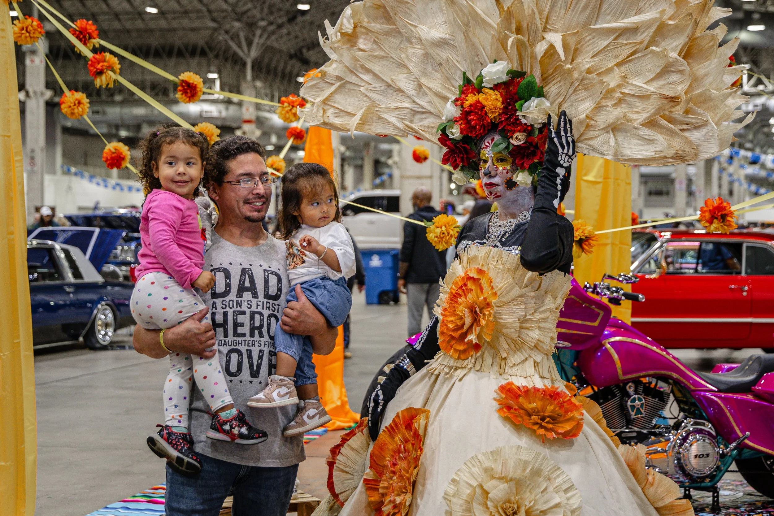 A man holding two young girls, posing next to a performer dressed in traditional Mexican attire with face paint and elaborate floral costume at an indoor event.