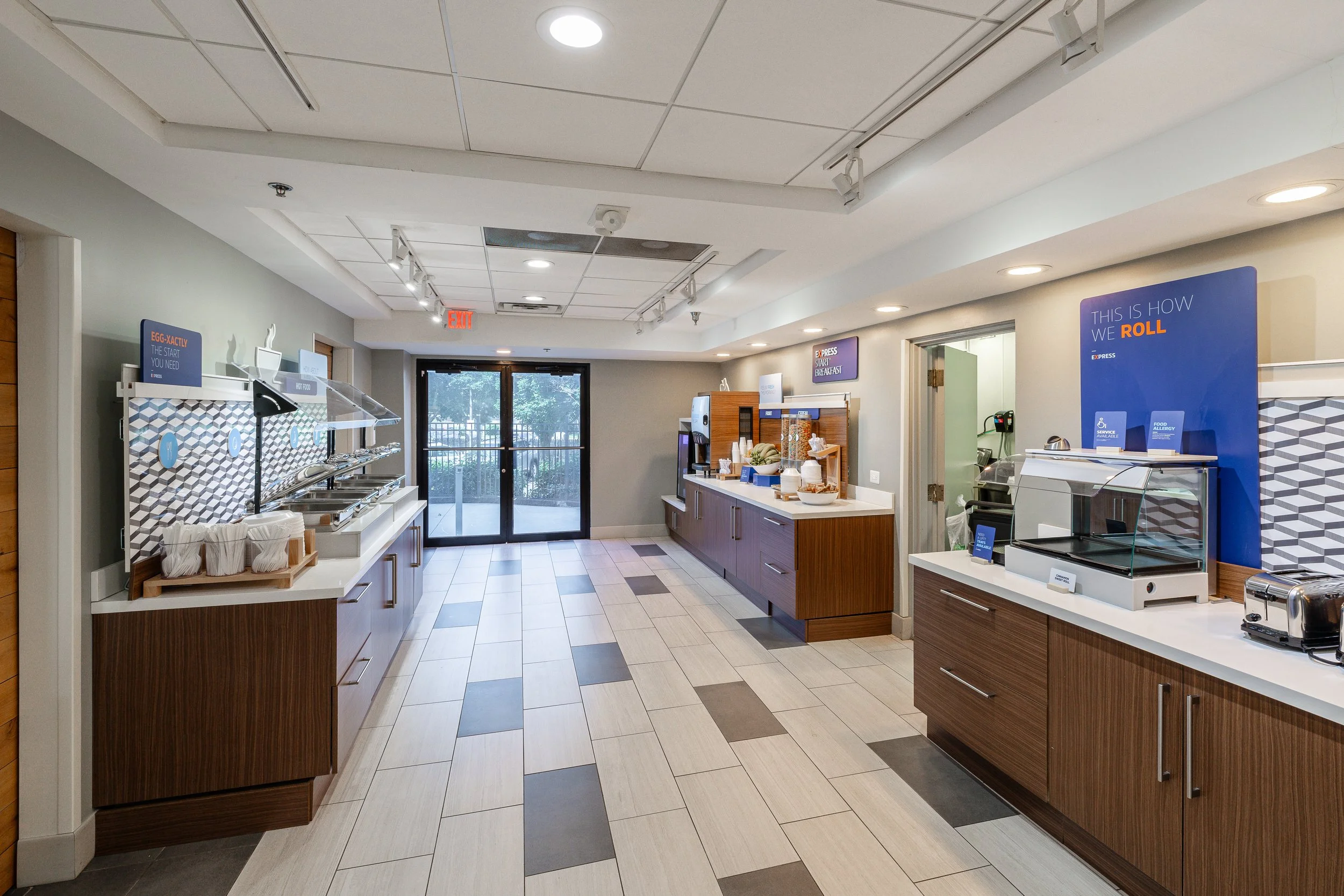 A breakfast buffet area with various stations, including a place for eggs and an array of breakfast items, with sliding glass doors at the back allowing natural light into the room.