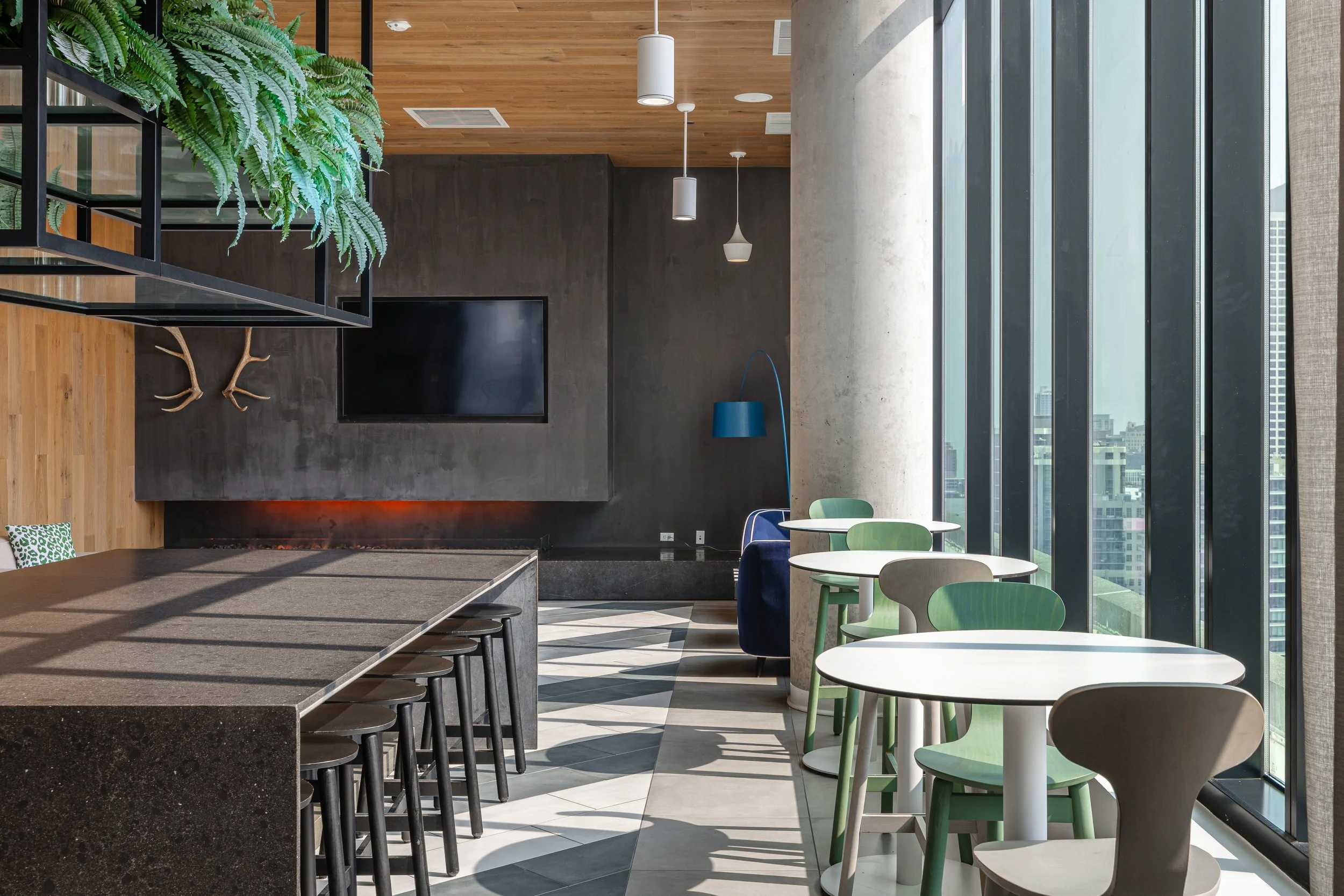 Modern cafe interior with a large window, green and white chairs, small round tables, a long dark bar counter, a flat-screen TV on a black wall, a blue arc floor lamp, a concrete column, wooden ceiling, hanging lights, and a staircase with decorative