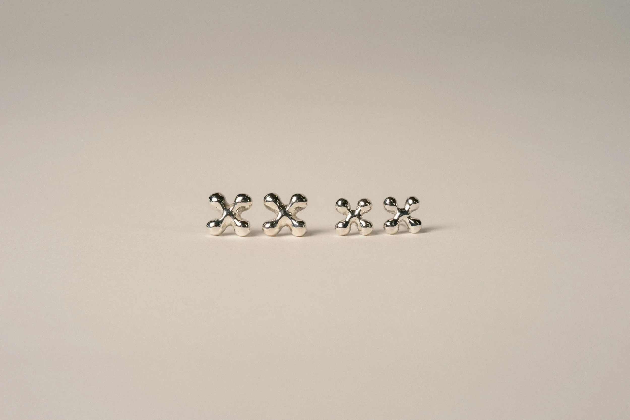 Four metallic X-shaped jewelry pieces arranged in a row on a plain, light-colored surface.