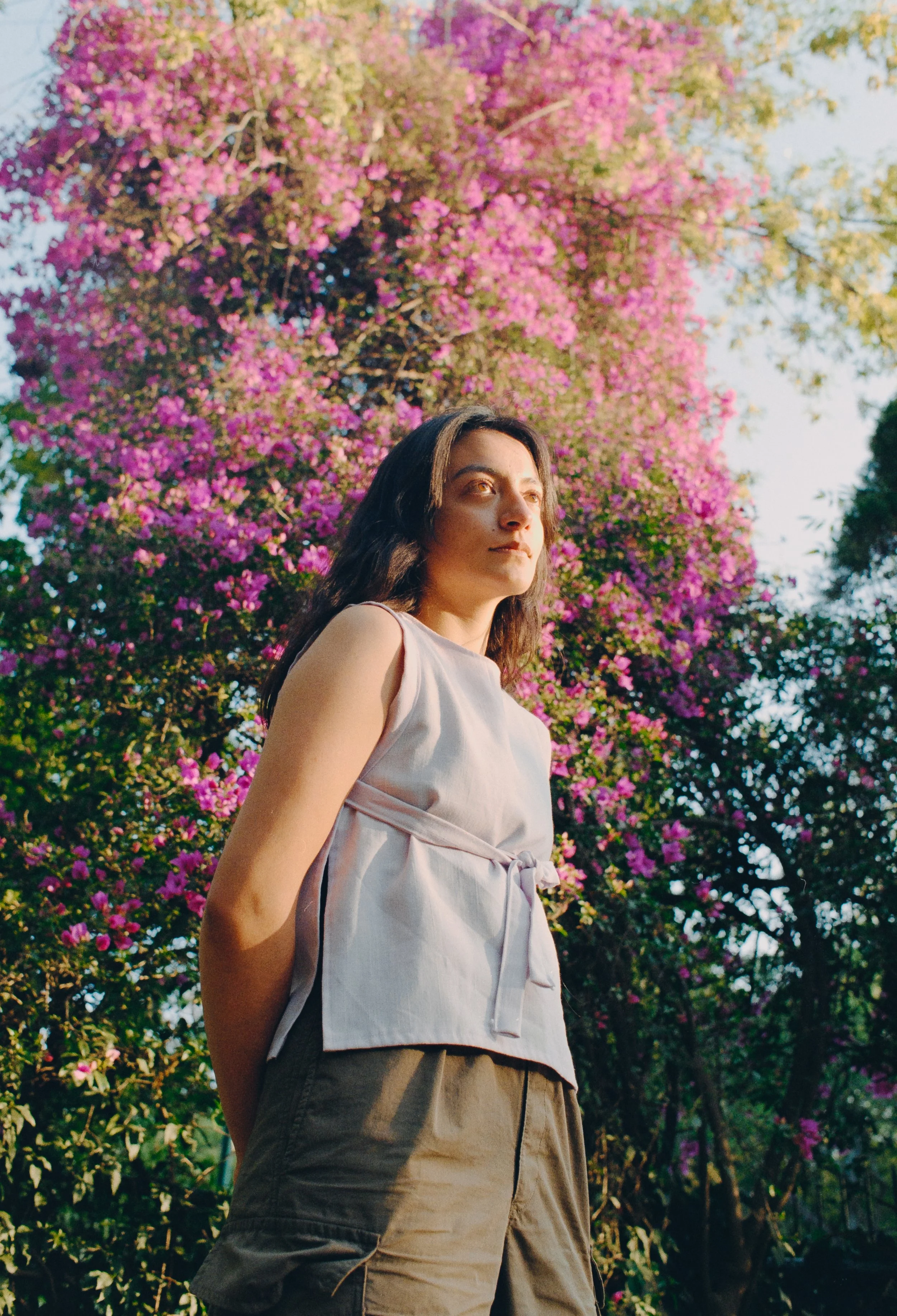 A woman standing outdoors near a blooming tree with pink flowers, looking into the distance.