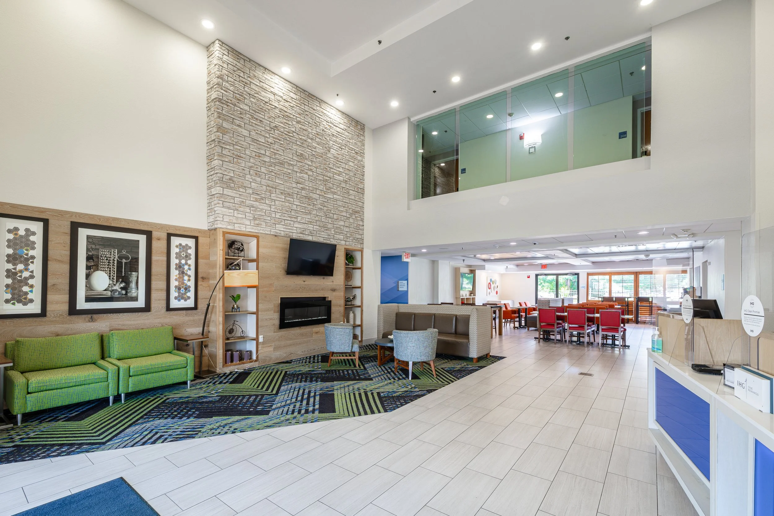 Interior of a modern lobby with a green sofa, light wood shelves, a TV, chairs, and tables, with a brick accent wall and large windows.