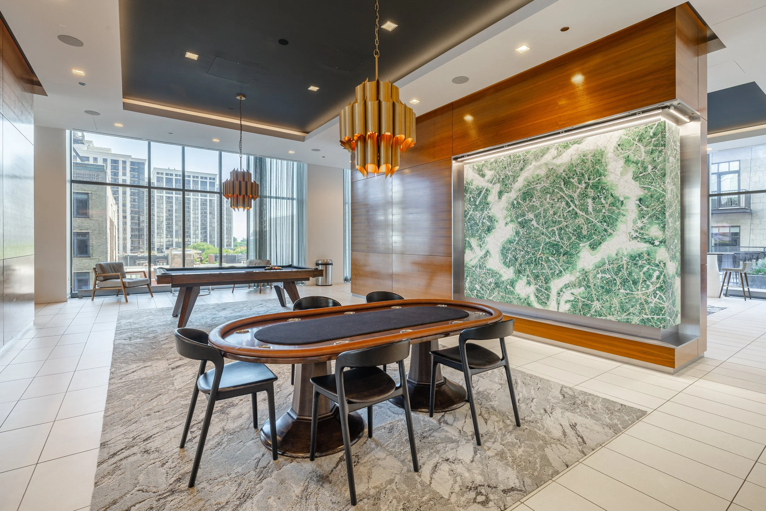 Modern game room with a poker table, a pool table, large window with city view, art piece resembling a green leaf, and contemporary lighting fixtures.