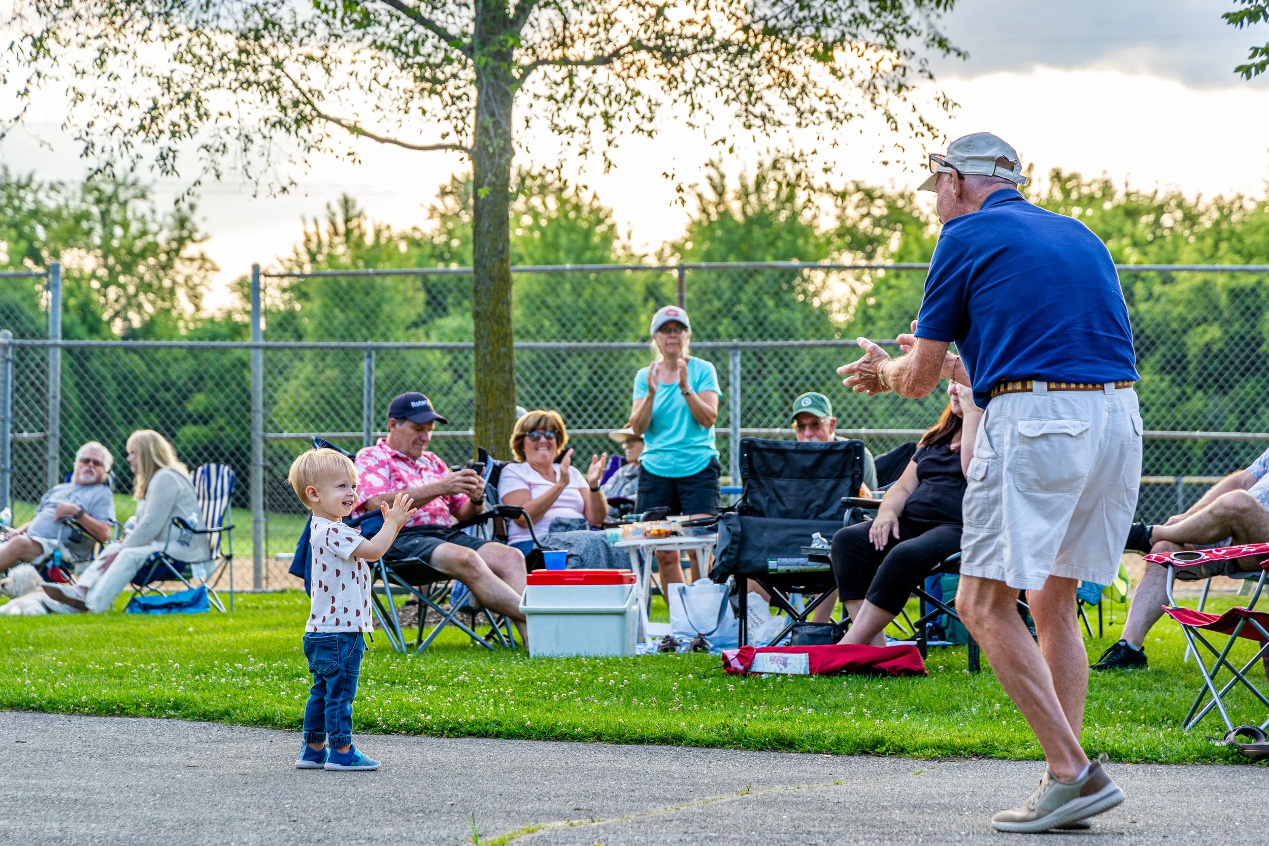 A young boy clapping and smiling at an outdoor gathering with several adults seated in lawn chairs and a man standing and speaking to the group, in a park near a baseball field fence, during sunset.