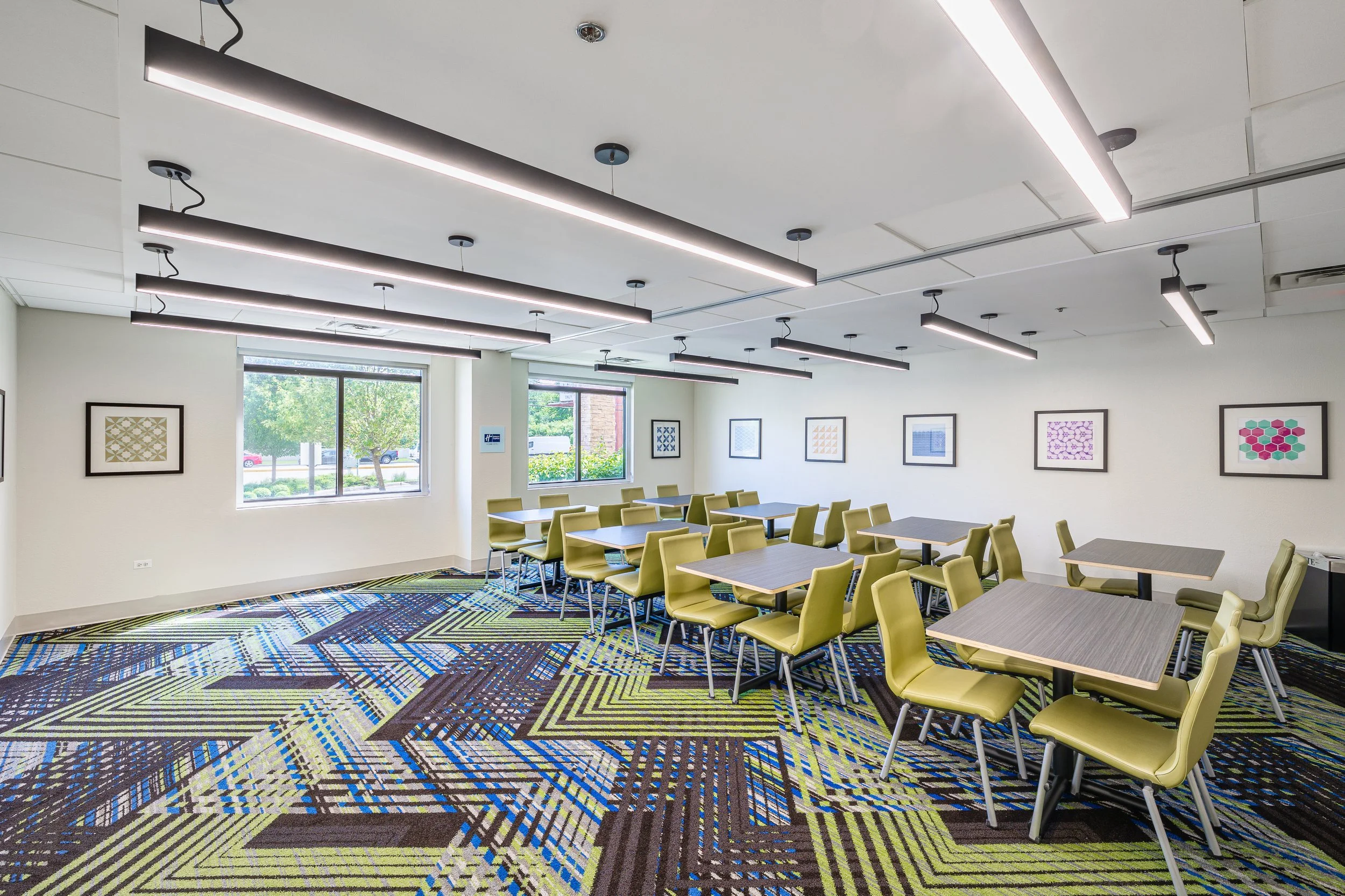A modern conference room with multiple green chairs and several tables. The room has large windows, framed artwork on the walls, and a colorful patterned carpet. There are contemporary ceiling lights.