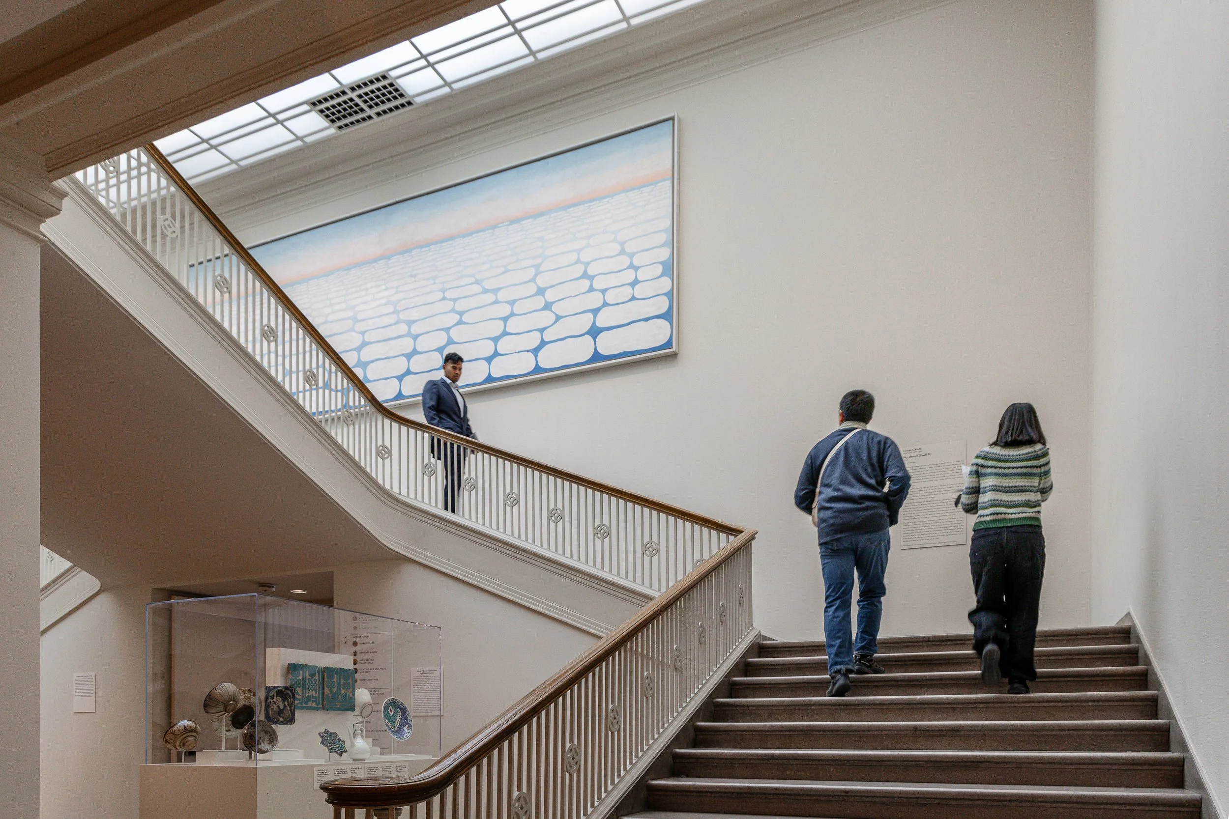 People walking up a staircase in an art gallery, with a large painting on the wall above them.