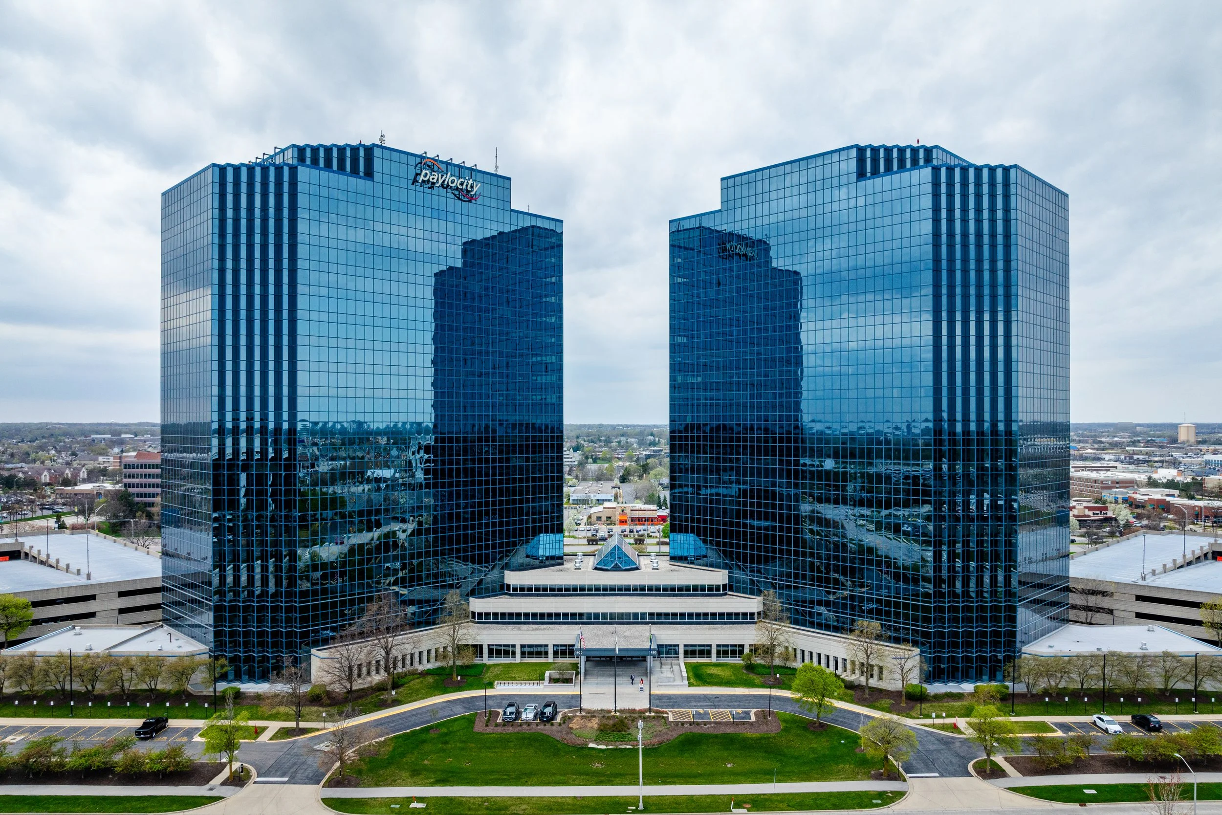 Two modern glass office towers in a corporate building complex with trees and parking lot in foreground, overcast sky in background.